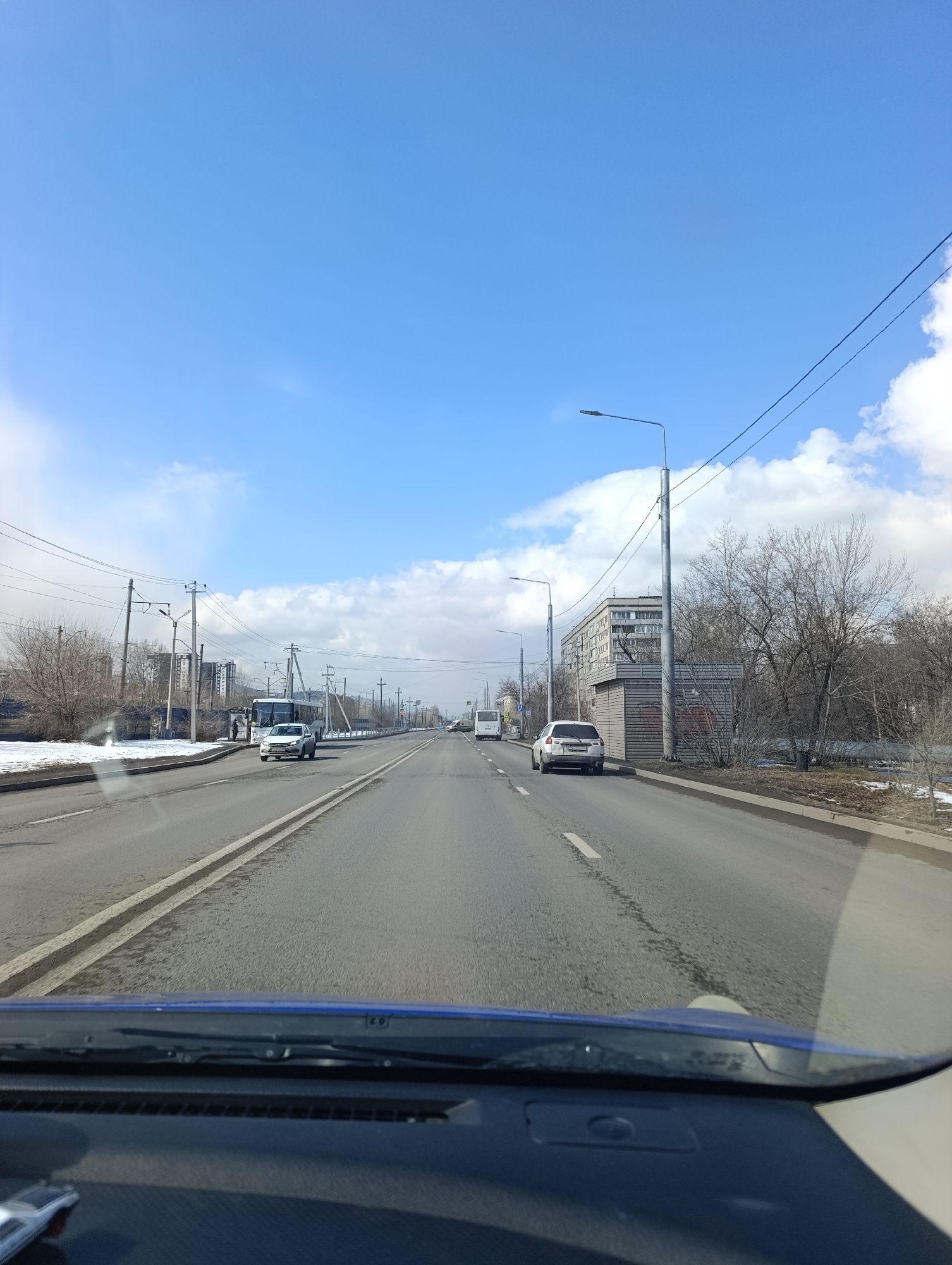 The image depicts a view from inside a moving vehicle on a multi-lane road in Krasnoyarsk, Russia. The road has lane markings and a median. The weather is sunny with a blue sky and scattered clouds. Along the side of the road are buildings, utility poles, trees without leaves, and some patches of snow. Other vehicles, including a bus and a white car, are visible on the road. The vehicle is moving and the dashboard is partially visible at the bottom of the frame.