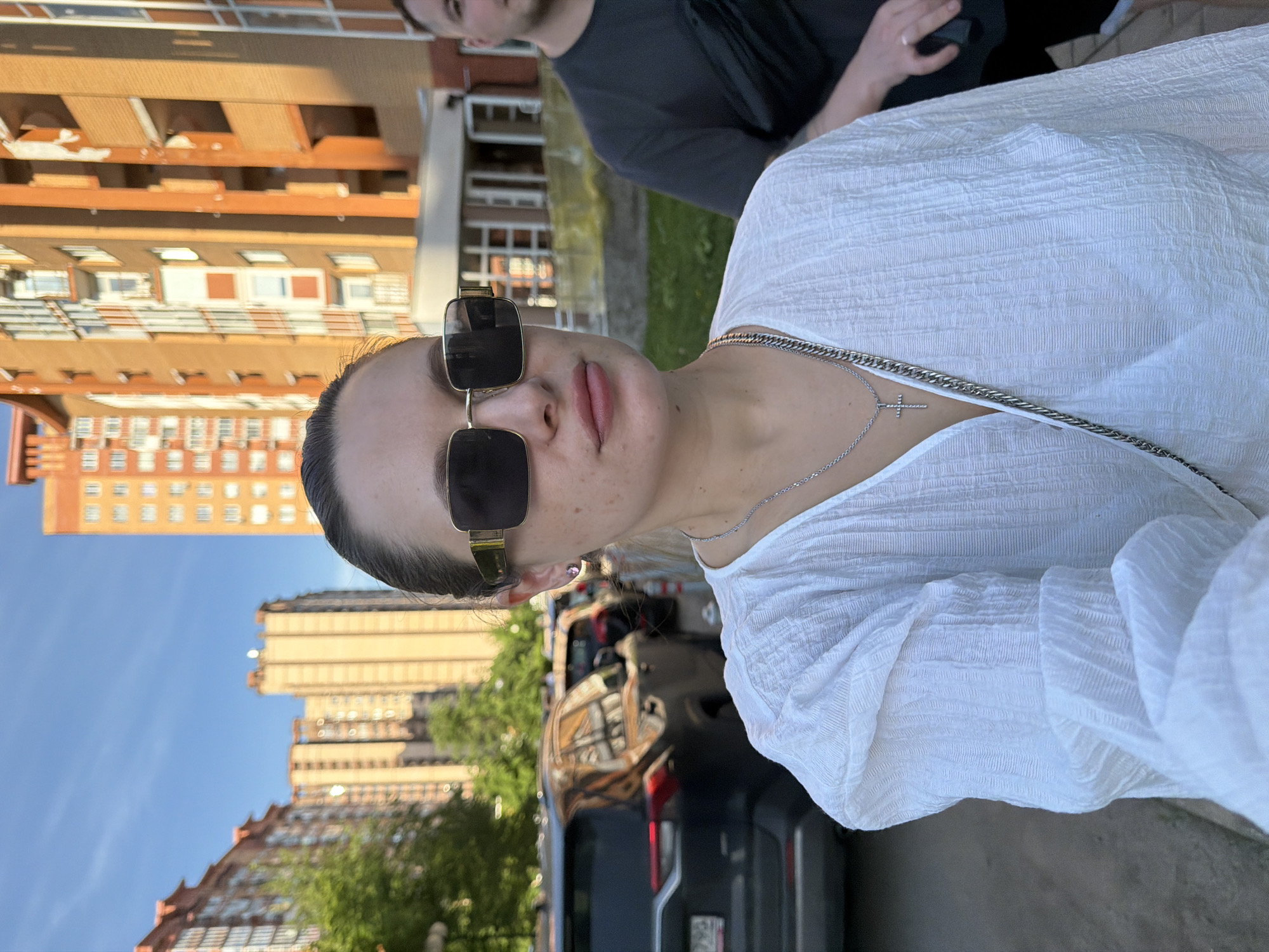 A young adult woman is positioned centrally in the foreground, taking a close-up selfie or being photographed. She has fair skin with visible freckles and her dark hair is pulled back. She wears large, rectangular, gold-framed sunglasses with dark lenses. Her lips are full and slightly parted. She is dressed in a loose-fitting, white, textured, v-neck blouse with gathered sleeves. Around her neck, she wears a silver chain necklace with a small, sparkling cross pendant, and a second, thicker silver chain. A purple stud earring is visible in her right ear. A silver chain from a bag or purse is draped over her left shoulder.
Behind her, on the right side of the frame, a young adult man is partially visible. He has short dark hair and wears a dark grey t-shirt. A black fanny pack or waist bag is worn diagonally across his chest. A wedding band is visible on his left ring finger.
The scene is an urban street in Odintsovo, Russia, under clear blue skies with bright daylight. In the midground to background, several multi-story modern apartment buildings are visible. They feature facades in varying shades of orange and brown brick or cladding, with numerous windows and balconies. Trees and green patches of grass are scattered between the buildings and alongside the street. On the left side of the frame, several cars are parked, including a dark grey SUV with a partially visible Russian license plate (ending in 90). The road surface is asphalt, and concrete elements of a sidewalk or curb are present.