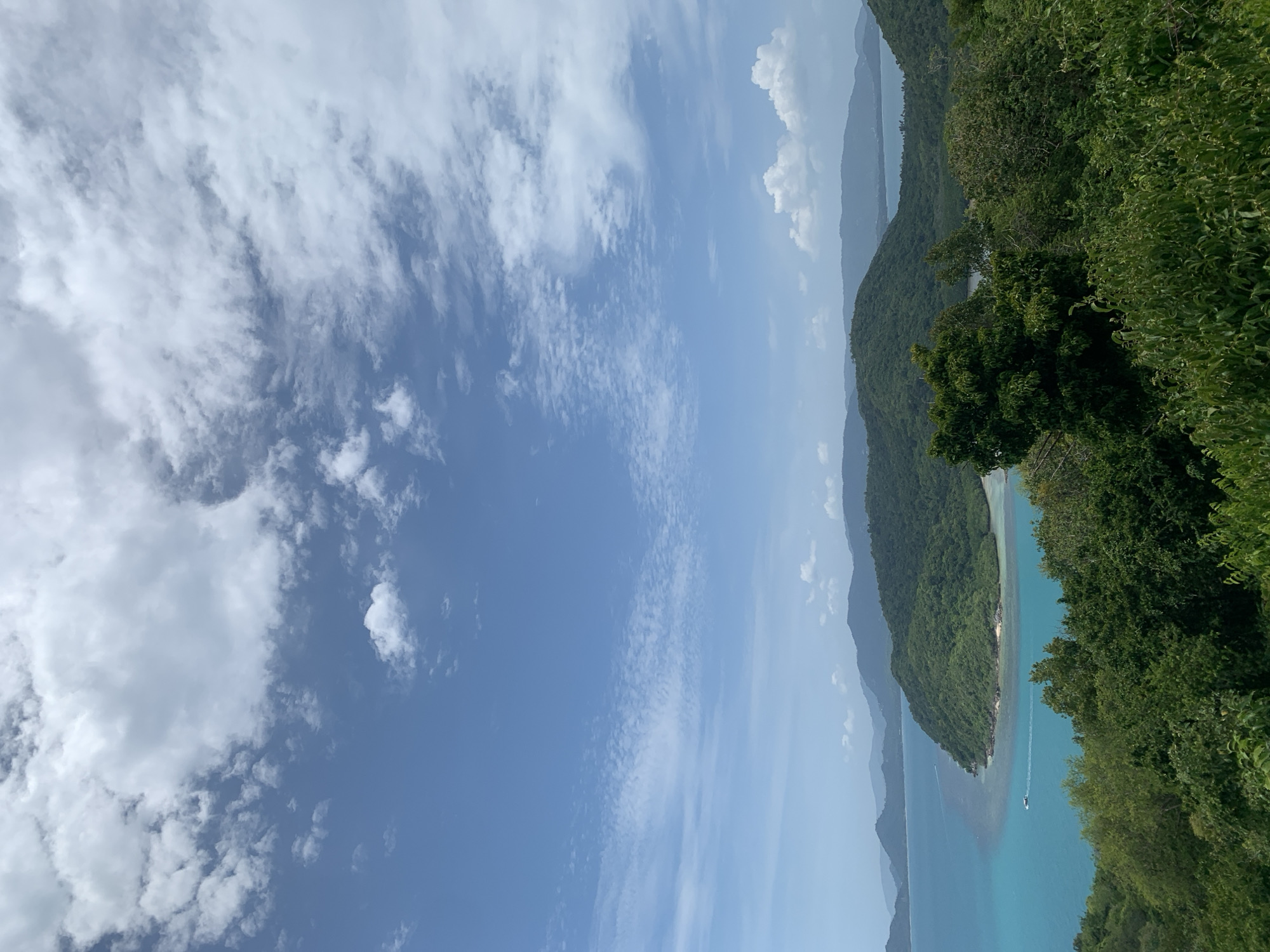 A high-angle, wide shot captures a panoramic view of a lush, green mountainous coastline meeting turquoise waters under a partly cloudy sky. In the foreground, dense foliage and trees create a natural frame. The middle ground showcases rolling, verdant hills that slope down to a vibrant, shallow bay. A small white boat is visible in the water. In the background, a series of hazy, blue mountains recede into the distance, and the ocean continues to stretch towards the horizon. The sky is a bright blue, dotted with fluffy white clouds, some of which are thin and wispy, while others are larger and more substantial. The overall scene depicts a serene tropical landscape.