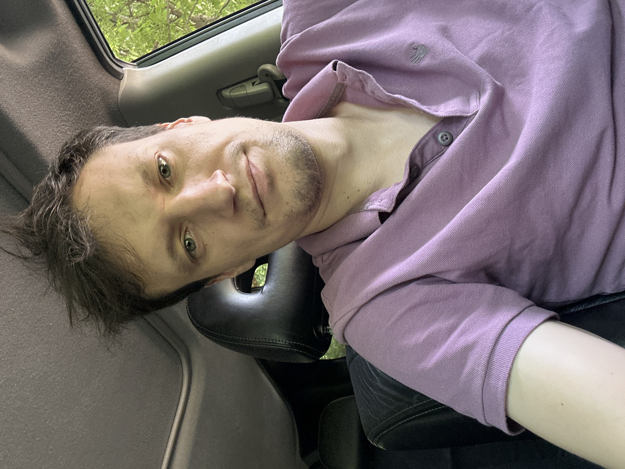 A man is sitting in the driver's seat of a car, taking a selfie. He has light skin, light brown hair, and green eyes. He is wearing a purple polo shirt. The car's interior is visible, including the headrest, sunroof, and car seat. Trees and foliage are visible through the car window, suggesting an outdoor location. The image does not contain any information indicating it was taken in Shatava, Ukraine.