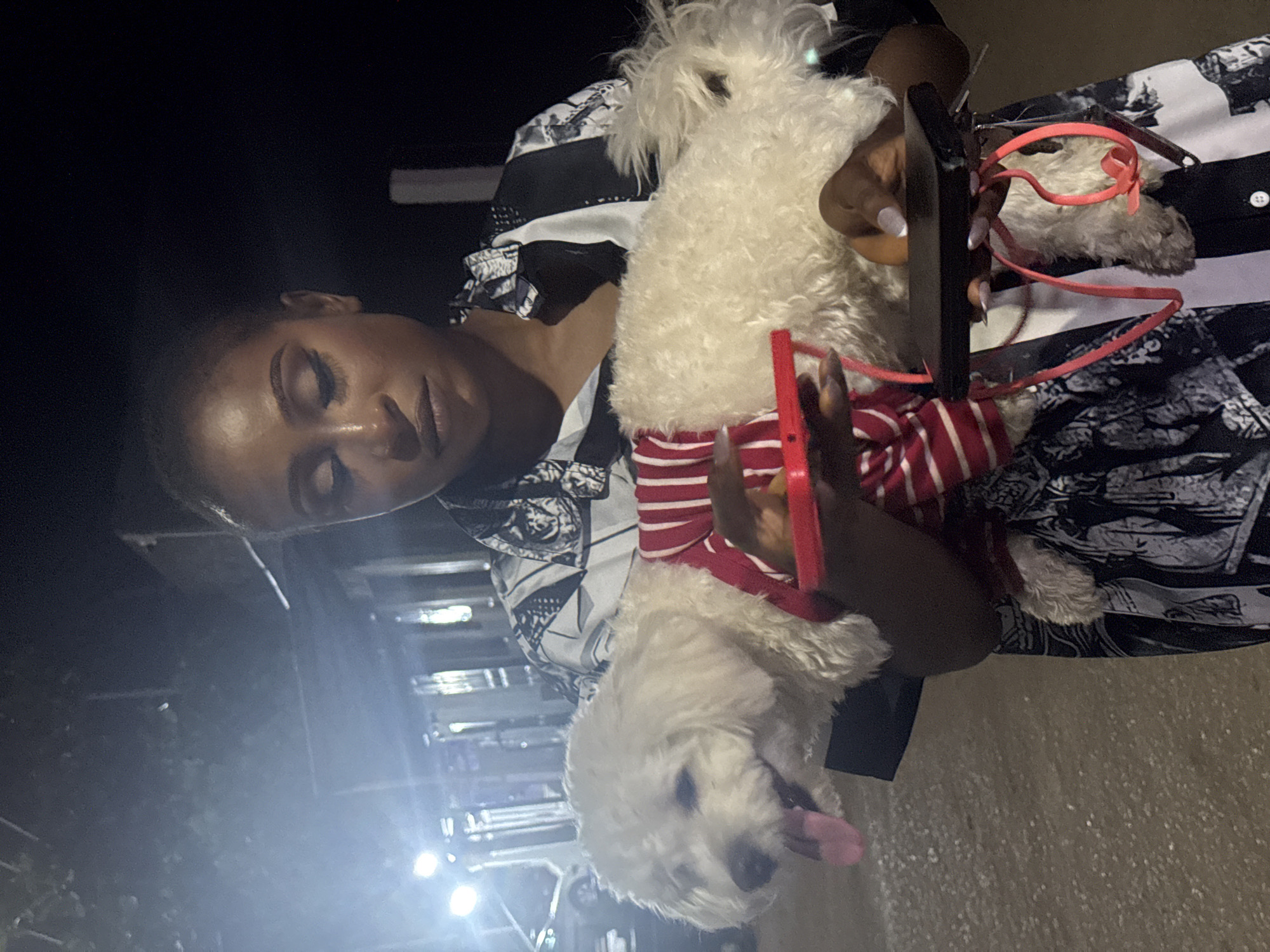 In Abuja, Nigeria, a woman with dark skin and short dark hair, wearing a black and white patterned shirt, holds a small white fluffy dog. The dog is wearing a red and white striped shirt and has its pink tongue sticking out. The woman is holding the dog with both hands, one hand under its belly and the other supporting its back, where a black electronic device with red straps is also being held. The background is dark and appears to be outdoors at night, with some blurry lights and structures suggesting a street or pathway. The ground beneath them is a light-colored gravel or sand.