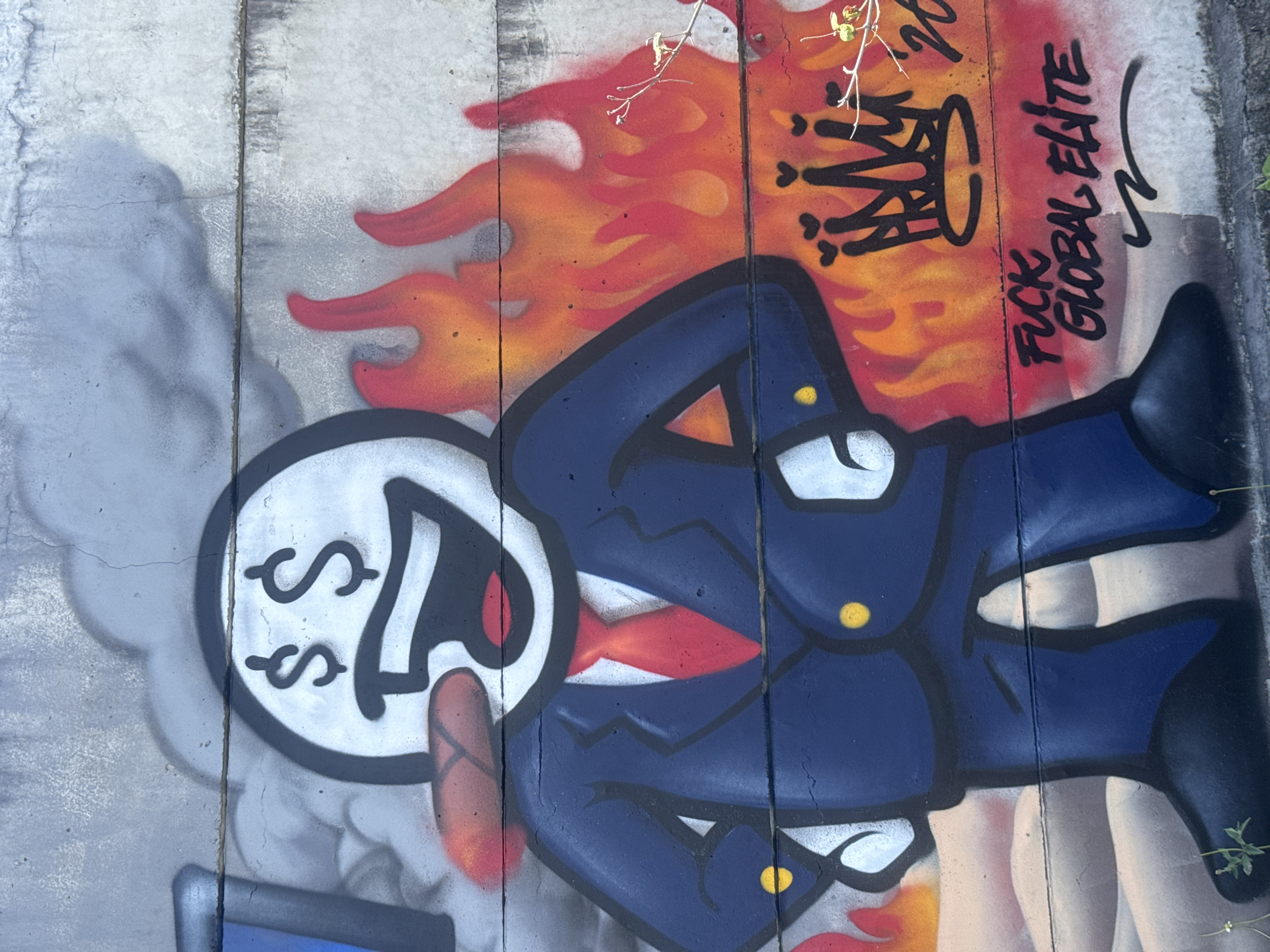 A colorful spray-painted mural adorns a concrete wall in Kuta Selatan, Indonesia. The central figure is a cartoonish character with a large, round head, sporting dollar signs for eyes and a wide, open mouth. The character is depicted wearing a dark blue suit with a bright red tie and yellow buttons. It holds a cigar in its mouth. Behind the character, flames in shades of red, orange, and yellow erupt. Above the flames and to the right of the character's head, a black crown with small hearts on its points is painted. Below the flames and to the right of the character's legs, the words "FUCK GLOBAL ELITE" are spray-painted in black. The overall scene is gritty and rebellious, with the concrete wall showing signs of age and weathering.