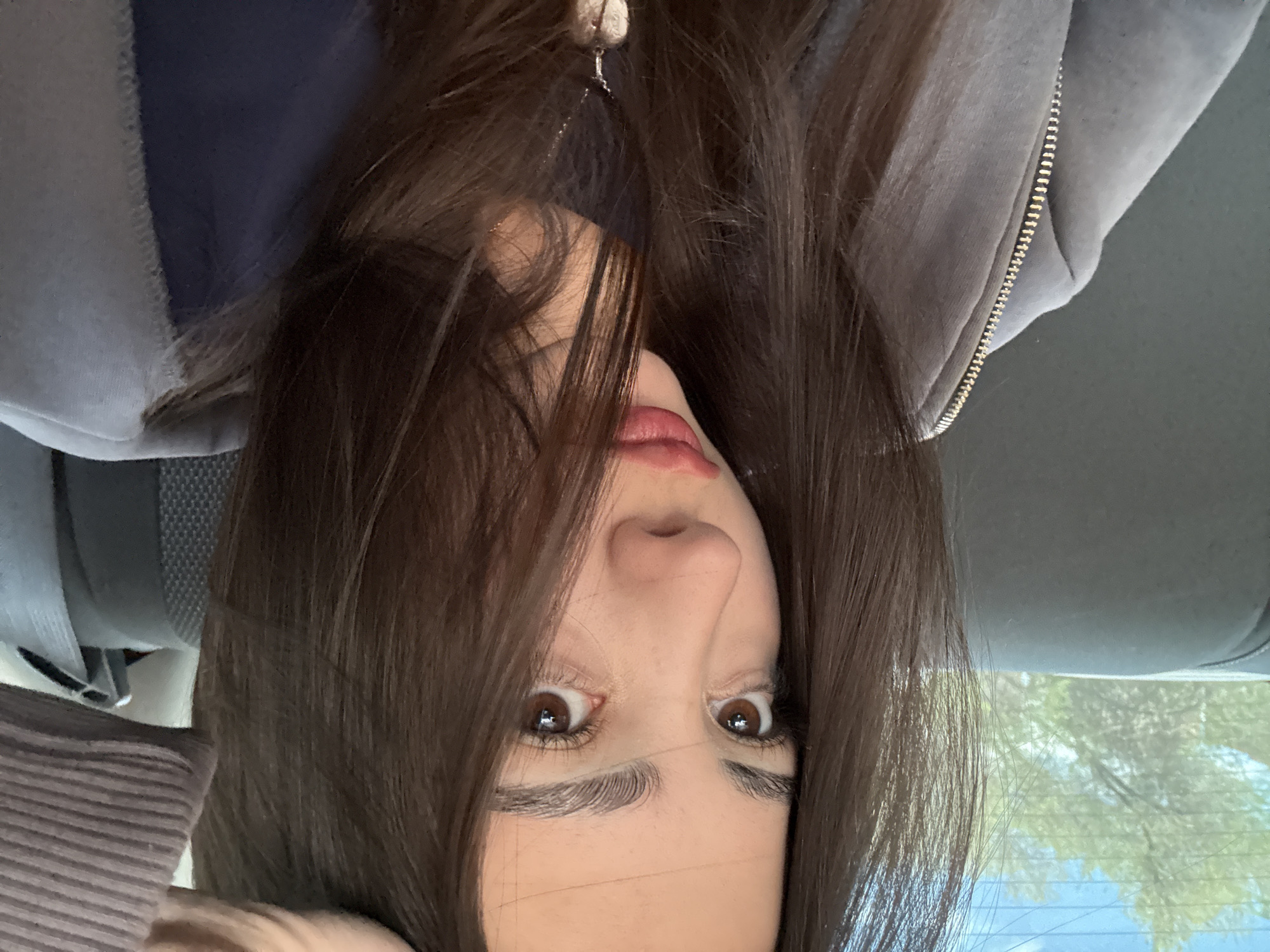 A young woman with long, dark brown hair is positioned upside down in what appears to be the back seat of a car. She has brown eyes and appears to be looking directly at the camera. She is wearing a grey hooded jacket with a zipper. A seatbelt is visible in the background. Through the car window, there is a glimpse of trees. It is not possible to confirm the specific location in Yerevan, Armenia, from this image alone.