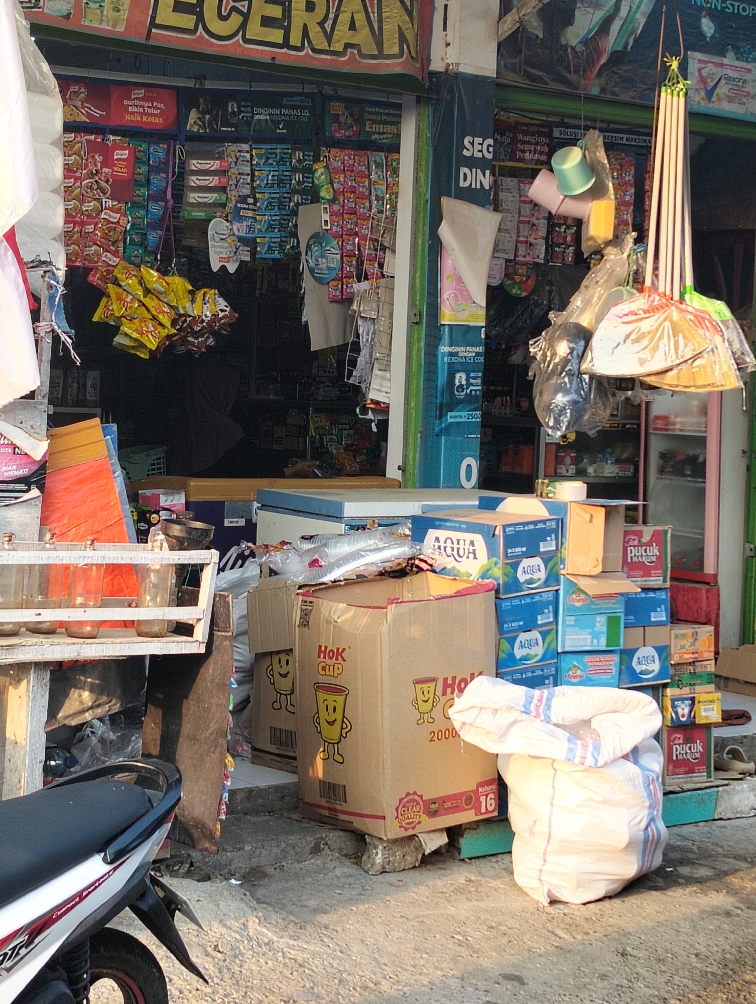 The image shows a street-side store in Banjarwati, Indonesia, with various items for sale displayed outside. In the foreground, a motorcycle is partially visible on the left. A white burlap sack, tied at the top and partially filled, sits on the concrete ground in the center. Behind the sack, several stacks of blue cardboard boxes, likely containing beverages, are arranged. To the left of the boxes, a large, light brown cardboard box with the text "HoK CUP" and an illustration of a smiling cup is prominent. Further to the left, a wooden shelf holds several glass jars with brown contents. The store itself is visible in the background, with shelves stocked with packaged goods. Hanging from the ceiling of the store's awning are several cleaning tools, including brooms. The overall scene suggests a busy, outdoor marketplace or a small convenience store.
