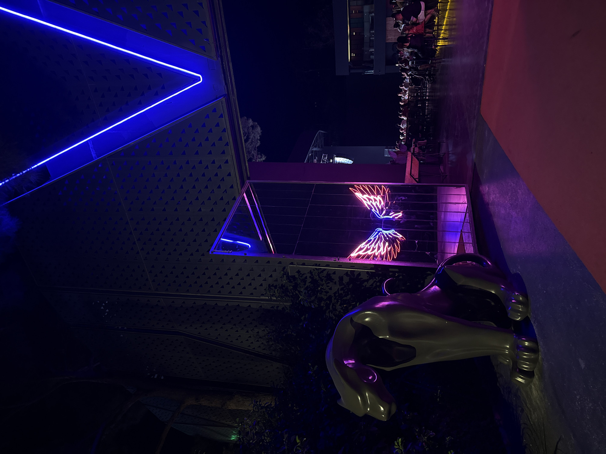 The image depicts an outdoor nighttime scene in Bodrum, Turkey, likely at a venue with an outdoor seating area. In the foreground to the left, a large, metallic sculpture of a stylized leopard or big cat sits, illuminated by purple and pink neon lights. To its right, a tall, rectangular mirrored structure stands, reflecting a neon sign of angel wings, glowing in pink and blue. Beyond this, the scene opens up to an outdoor seating area where numerous people are seated at tables. They are mostly silhouettes against the ambient lighting, with some individuals appearing to be engaged in conversation or dining. Above, a large, angular structure with a blue neon light tracing its outline is visible, suggesting a canopy or architectural feature of the venue. The overall atmosphere is modern and vibrant, with the neon lighting creating a dramatic and colorful ambiance against the dark night sky.