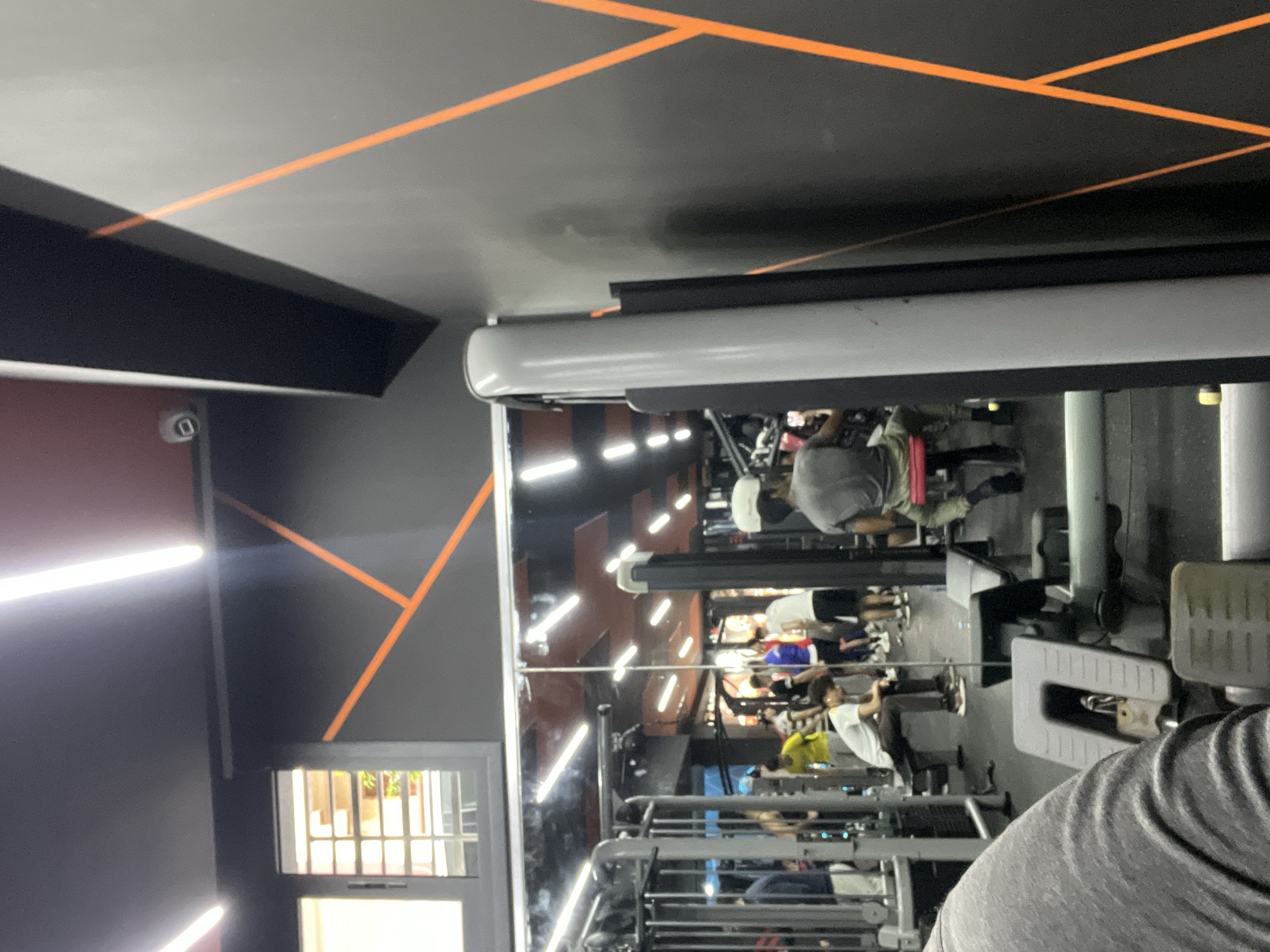 The image captures the interior of a modern gym or fitness center, viewed from a low perspective, likely from a seated position on a piece of equipment. The shoulder and upper arm of the person taking the photograph are visible in the lower right foreground.
The gym features dark grey or black walls and ceiling, strikingly decorated with bright orange geometric lines that crisscross various surfaces. The space is brightly illuminated by long, white LED strip lights, visible on the walls and reflected in the shiny surfaces.
A dominant feature is a large mirror wall on the right, reflecting the activities within the gym and creating an illusion of expanded space. In the mirror's reflection, several individuals are seen engaged in exercise. One person is distinctly visible using a rowing machine, while others are reflected at various weightlifting or cardio stations.
On the left side of the room, a window with security bars offers a view of some greenery outside. A white security camera is mounted on a structure in the upper left portion of the image. The setting suggests an active, well-equipped fitness environment. There is no legible text visible in the image.