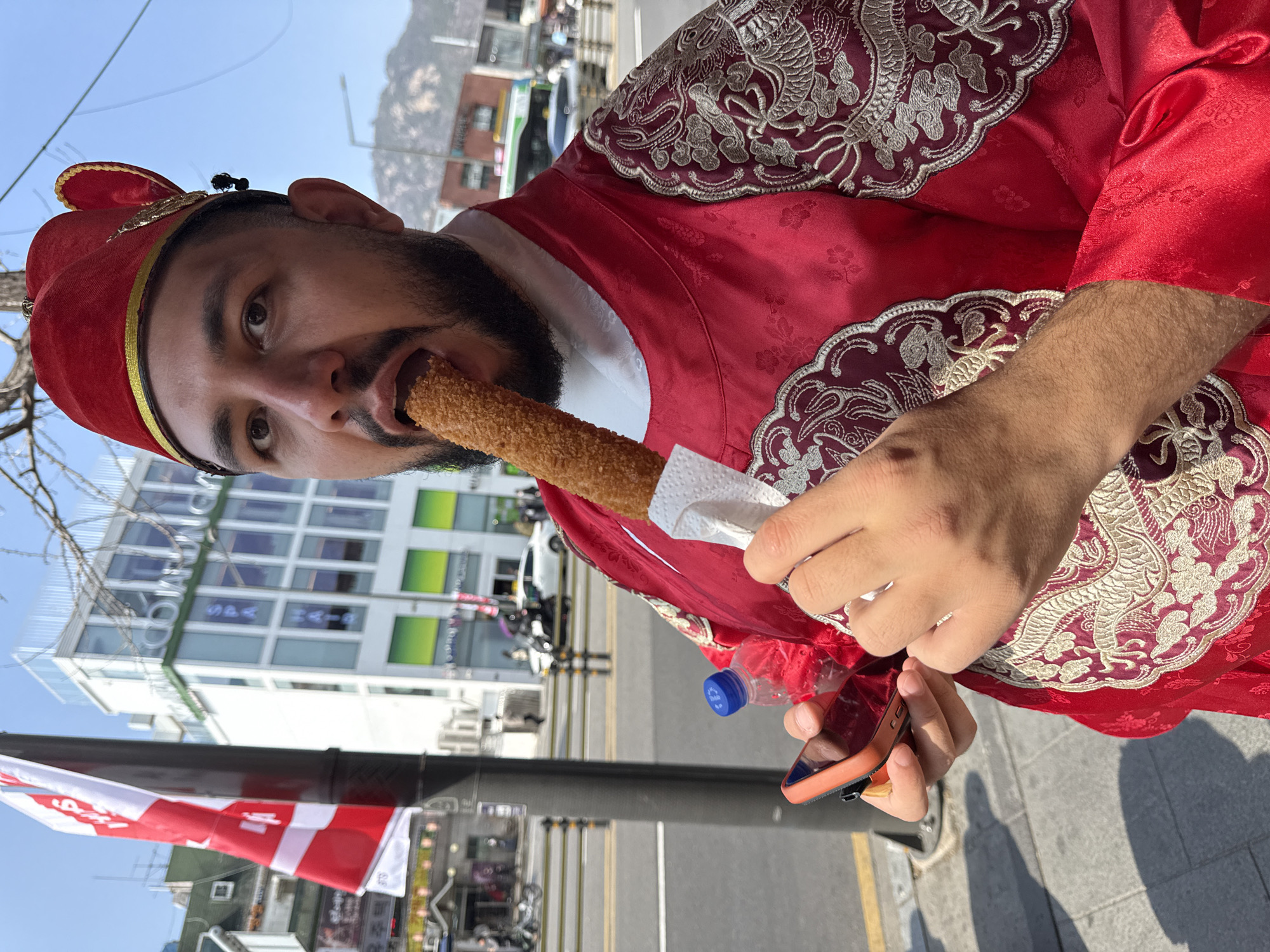 A male subject, with dark hair, a beard, and a mustache, is captured in a medium-shot on a sidewalk in Seoul, South Korea. He is wearing a traditional red Korean royal court attire, including an embroidered robe with gold patterns on the shoulders and a matching red cap. The subject is actively consuming a golden-brown, breaded fried food item, held in a white paper napkin, which is partially inserted into his open mouth. In his left hand, he holds a smartphone with an orange case and a small, clear plastic water bottle with a blue cap. His gaze is directed towards the viewer.
The background depicts a vibrant urban street scene. To the subject's left, a black utility pole supports a red and white flag displaying partial Korean text. Across the street, a prominent multi-story white building with green accents features large windows and visible signage, including "CODIUM" and "SPA HAHA," which identifies the location as Seoul, likely in the Jongno-gu area. Traffic, including cars and motorcycles, is visible on the asphalt road, with pedestrians in the distance. A distinct mountain range is visible in the far background under a clear blue sky. The immediate foreground consists of a grey concrete sidewalk.