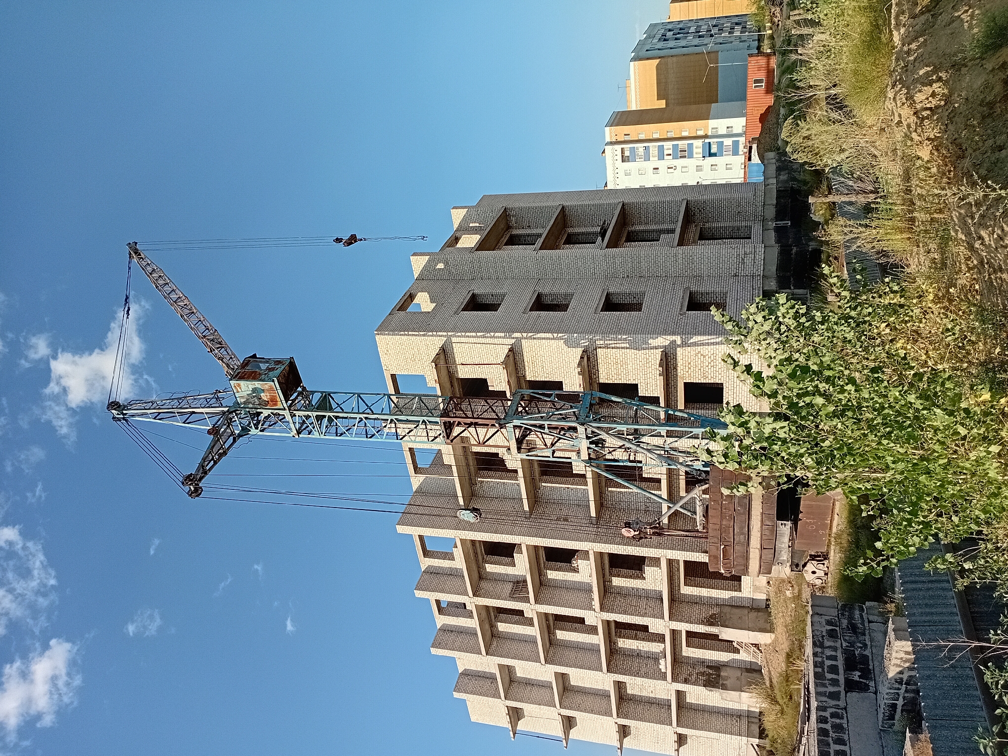 An outdoor, low-angle, full shot captures a construction site in Semey, Kazakhstan, under a clear blue sky dotted with sparse white clouds. In the foreground, a large, industrial tower crane, painted teal and rust, stands tall, its boom extending to the right. The crane's cabin is visible at the top of the main tower, and a hook hangs from the boom. To the left of the crane, a multi-story building under construction is partially visible, constructed from light-colored bricks with open, rectangular window and balcony openings. Heavy, dark metal counterweights are stacked at the base of the crane. The ground is covered with dry grass, dirt, and scattered construction materials, including concrete blocks. In the background, several finished apartment buildings with various colored facades are visible, including one predominantly blue and white, another orange, and one with a light beige exterior. A shipping container, painted orange, is also present behind the unfinished building.