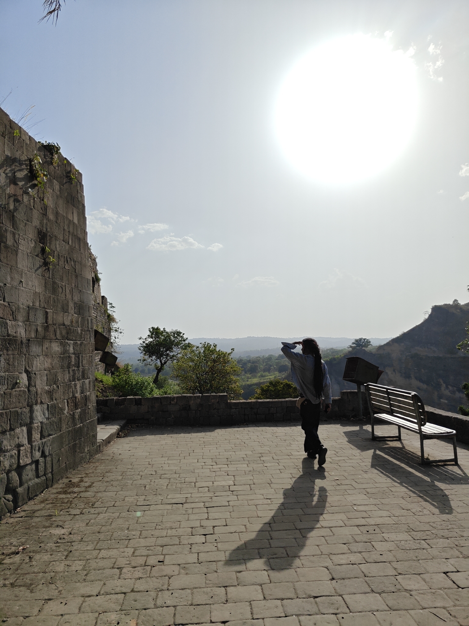 The image shows a person standing on a brick-paved terrace or platform in Kaswara, India, shielding their eyes and looking out towards a landscape.
The person is wearing dark pants and a light-colored, patterned long-sleeved shirt. Their hair is long. The sun is very bright in the sky, positioned to the right of the subject.
To the left of the person is a tall, aged stone wall. To their right, near the edge of the terrace, is a park bench and a small metal bin mounted on a pole.
The landscape visible beyond the terrace consists of rolling hills and trees. The overall atmosphere appears to be sunny and possibly hot.
