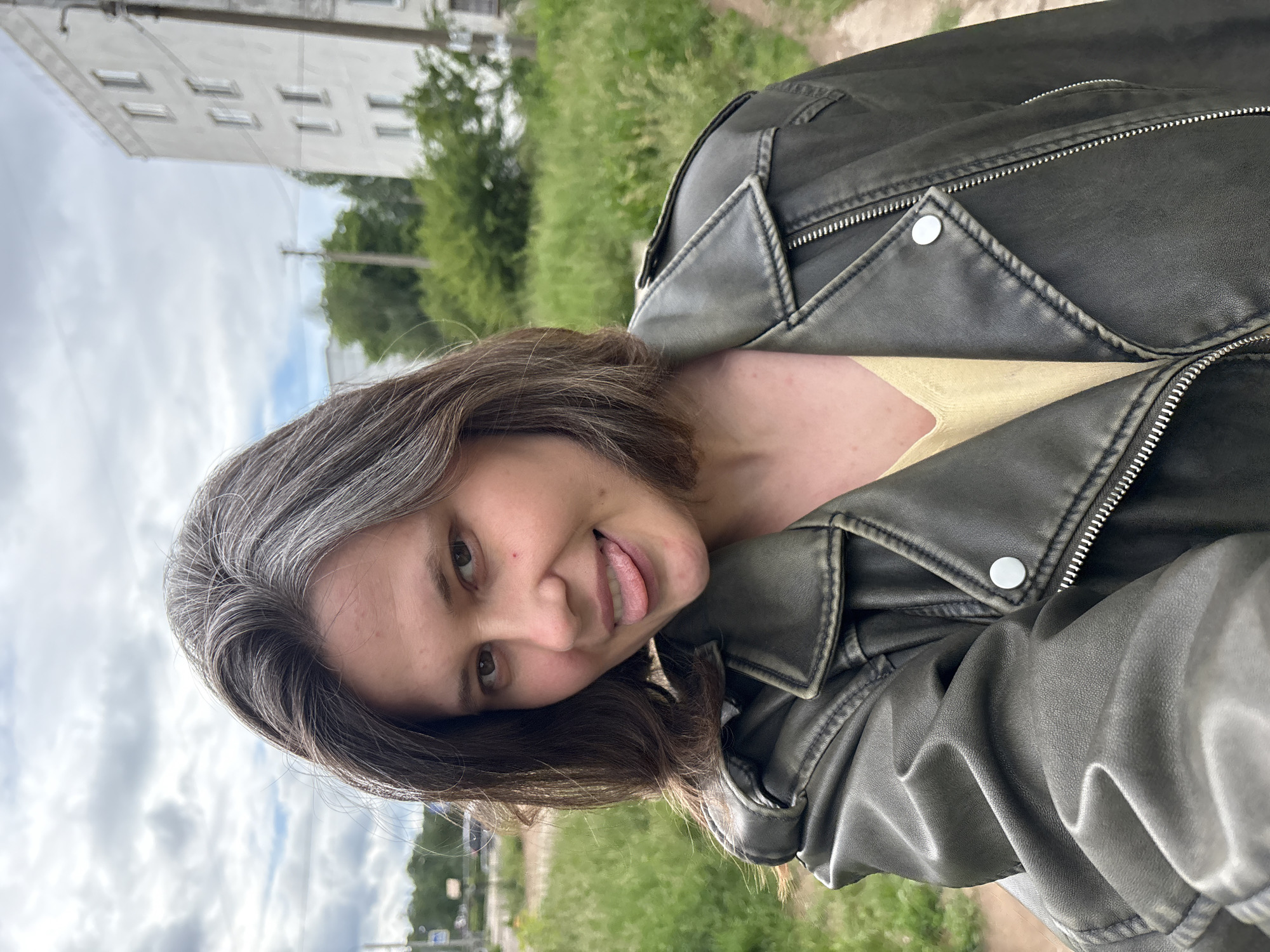 A casual, outdoor, half-body portrait features a young woman with a playful expression, sticking her tongue out at the camera. Her head is tilted, and the image is oriented sideways. She has medium-length dark brown hair with noticeable gray streaks, particularly around her temples and part. Her eyes are dark.
She is wearing a dark grey or faded black leather-style jacket with prominent silver zippers and snap buttons, giving it a somewhat distressed look. A light yellow top is visible underneath the jacket.
The background depicts an urban-natural mix. In the upper left (when the image is correctly oriented), a light-colored, multi-story building with numerous rectangular windows is visible under an overcast sky. Below the building and extending across the mid-ground is a lush green landscape with trees and abundant grass, suggesting a park or green belt. A dirt path runs along the right side of the frame. A utility pole and overhead wires are faintly visible in the distance. The sky is cloudy, suggesting a bright but not sunny day. The location context is Chaikovsky, Russia. There is no visible text in the image.