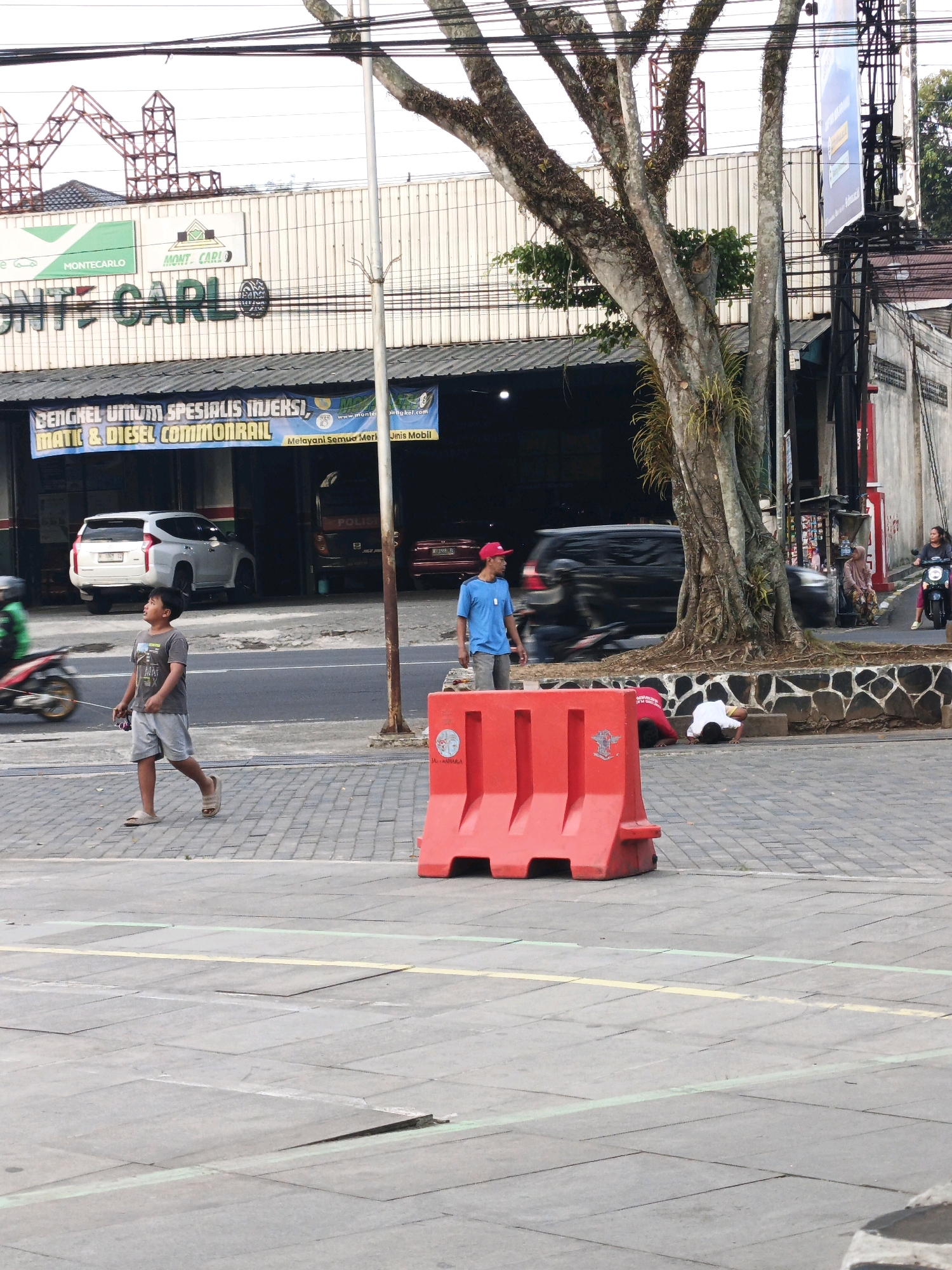 In a city street scene, a young boy in a grey t-shirt and shorts walks on a brick-paved sidewalk, holding a string. He is looking up and to his left. To his right, a large red plastic traffic barrier is positioned on the sidewalk. Behind him, a white SUV is parked in front of a building with large banners that advertise automotive services. Further down the street, several people are visible, including a man in a blue shirt and red cap standing near the barrier, two individuals kneeling on the ground behind the barrier, and a woman riding a motorcycle. A motorcycle is also seen speeding by in the background. The environment appears to be a commercial area with overhead power lines and a large tree with exposed roots on the right side of the frame.