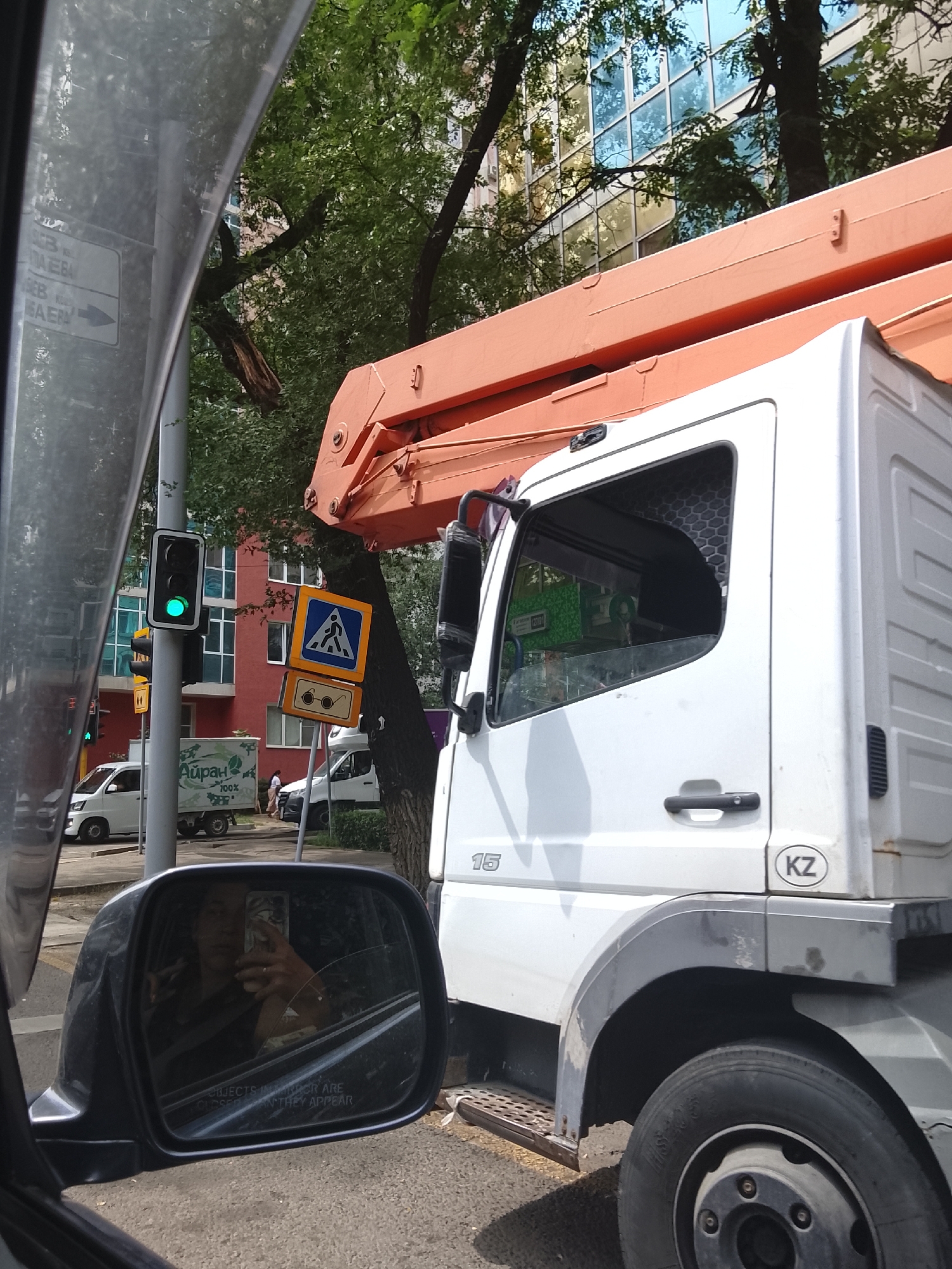 The image shows a street scene in Almaty, Kazakhstan. The foreground features the driver's side mirror of a vehicle, reflecting the driver taking a selfie. To the right of the mirror is the side of a white truck with an orange construction boom. The truck has a "KZ" sticker on its side, indicating it is registered in Kazakhstan. In the background, there is a traffic light showing green, a pedestrian crossing sign, and a building with a glass facade. A white truck with the logo "Айран 100%" is visible in the middle ground. Trees with green foliage are scattered throughout the scene. The overall impression is that of a busy urban environment on a bright, sunny day.