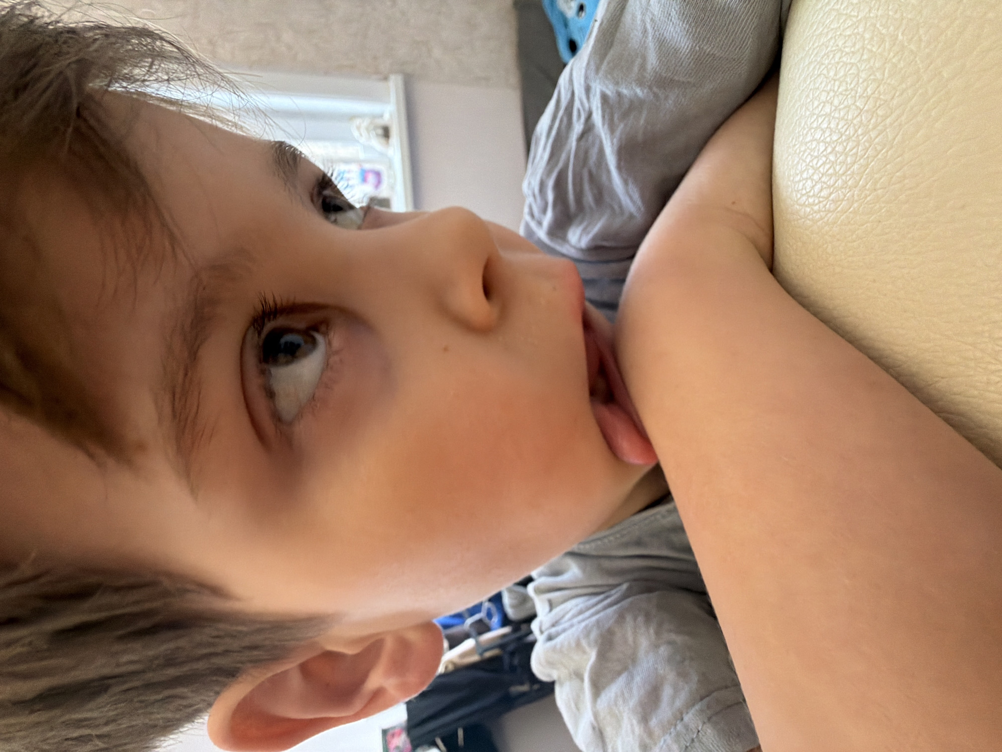 A young child, likely male, is captured in a close-up, tilted shot. The child is fair-skinned with brown hair and eyes. His tongue is extended, touching his arm, which is bent and resting on a light-colored, textured surface, possibly a leather or vinyl couch. He is wearing a light grey, short-sleeved shirt. The background is blurred, showing a window with white blinds or trim and other indistinct objects. There is no information to determine the location in the city of Perm, Russia.