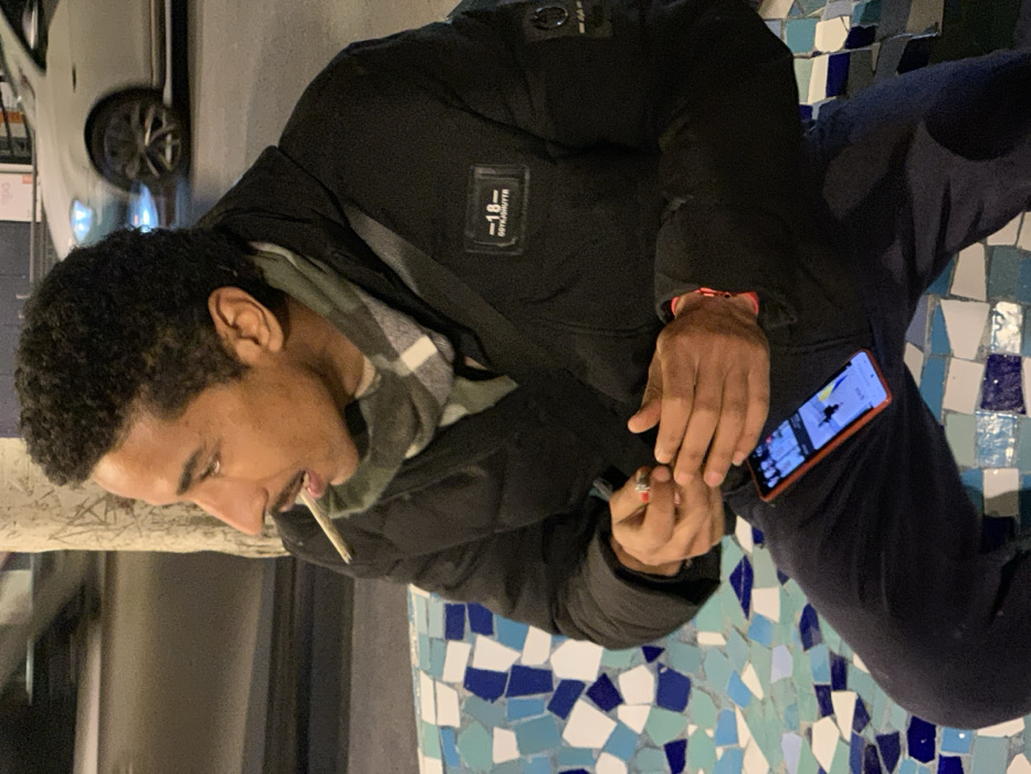 An individual, seemingly male, sits on a mosaic-tiled surface. He is dressed in a black puffer jacket, dark pants, and a plaid scarf. He is holding a lit cigarette or joint in his mouth and in one hand, using a lighter with the other. Next to his leg is a smartphone with a bright screen showing a snowy mountain scene. Part of a white car is visible in the background.