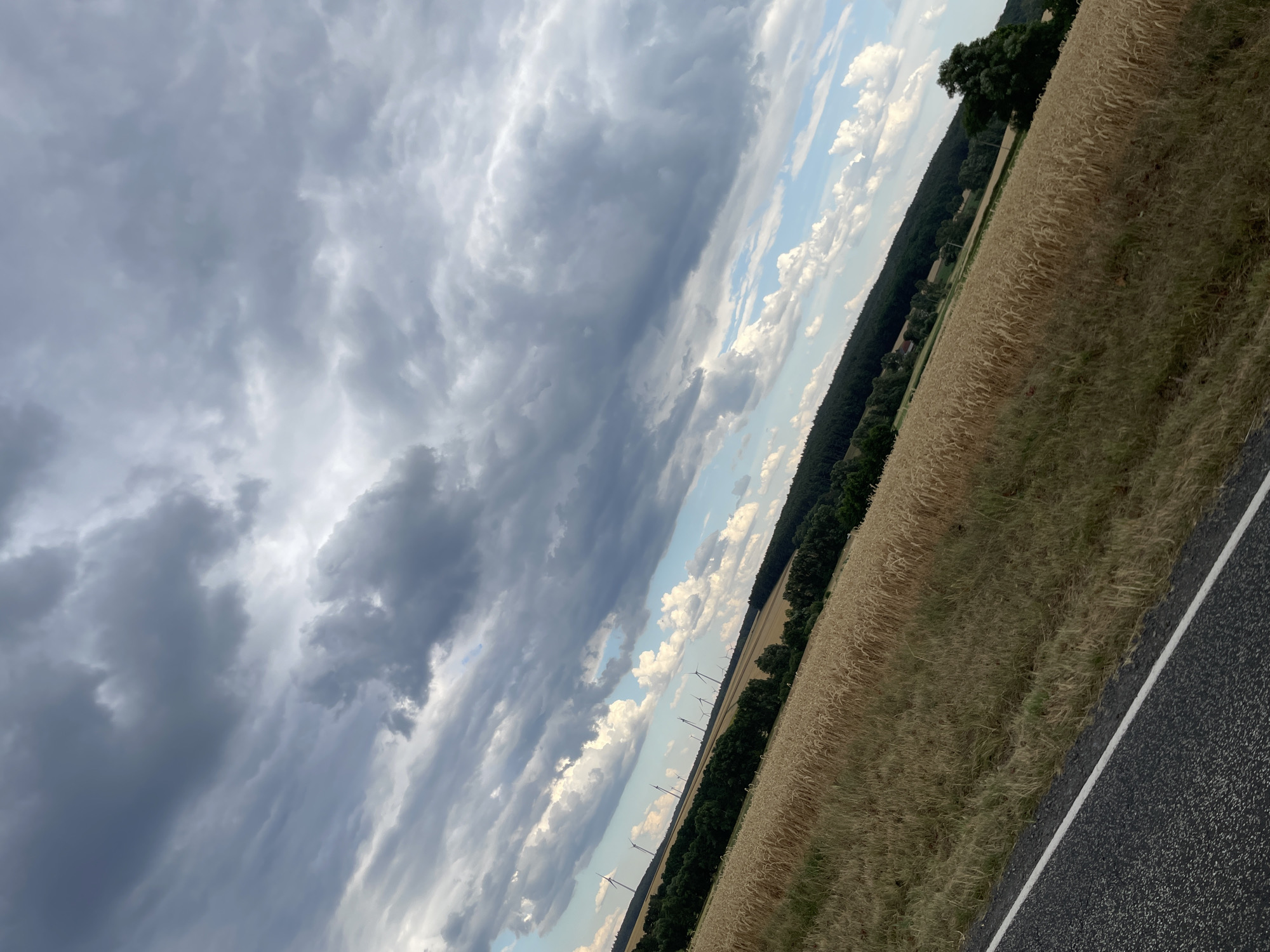 This image shows a landscape view of a field of wheat with a dark grey asphalt road in the foreground. The field extends into the distance and is bordered by a dense line of dark green trees. Beyond the trees, several wind turbines are visible against a sky filled with dramatic, dark grey clouds interspersed with patches of blue sky and white clouds. The overall scene suggests a rural setting, likely in the vicinity of Holzburg, Germany, characterized by agricultural land and renewable energy infrastructure. There are no people or animals visible in the image. The lighting suggests it is late afternoon or early evening, with the heavy cloud cover creating a moody atmosphere.