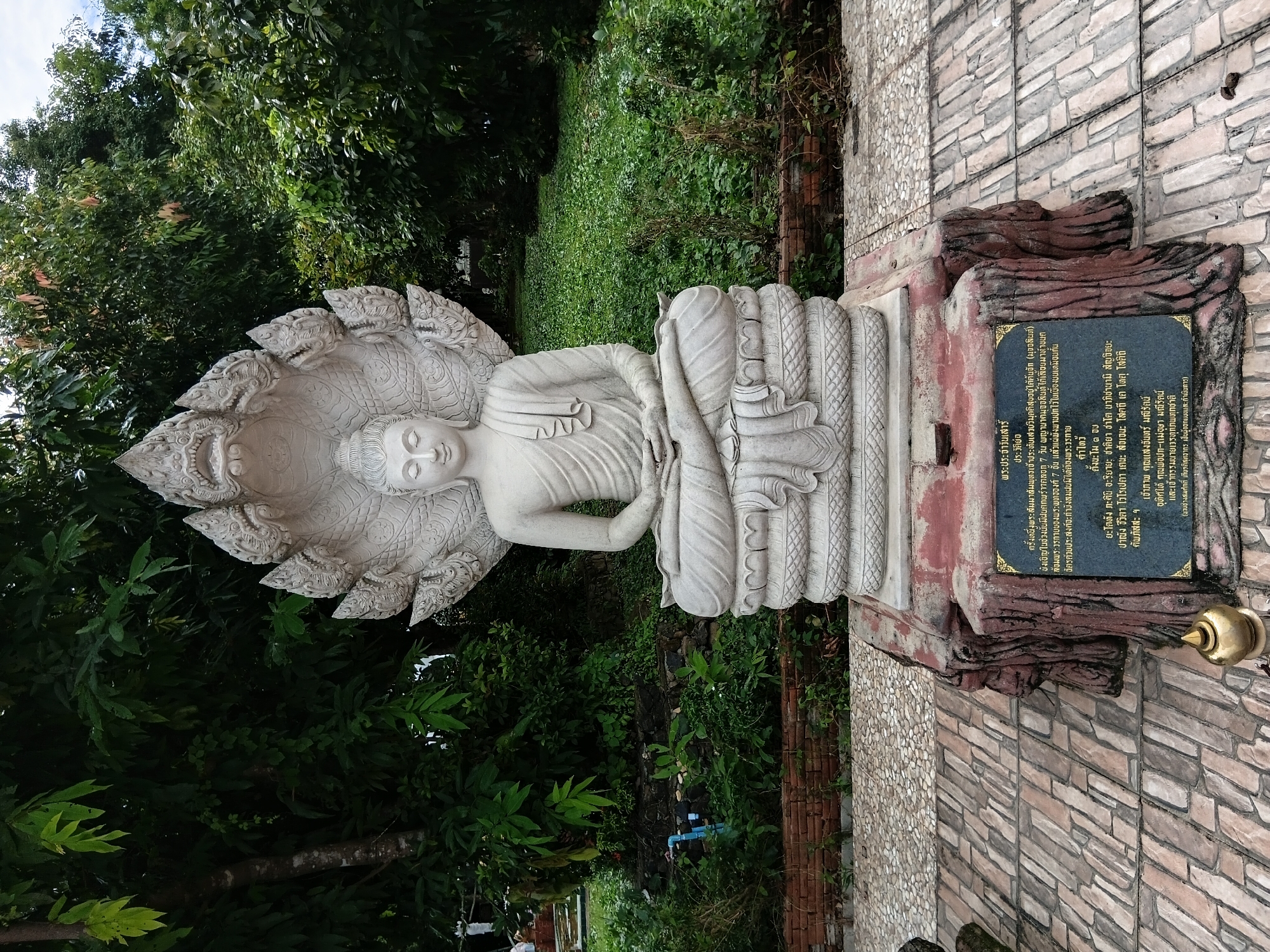 The image displays a stone statue of Buddha seated in meditation, with a multi-headed Naga serpent providing a canopy over the Buddha's head. The statue is positioned on a decorative base made of what appears to be faux wood, and it stands on a paved area with a patterned stone walkway. Behind the statue, lush green trees and vegetation create a natural backdrop. A plaque with Thai inscriptions is affixed to the base of the statue, providing context or historical information. The scene appears to be outdoors in a temple or garden setting within Ban Huai Kit, Thailand.