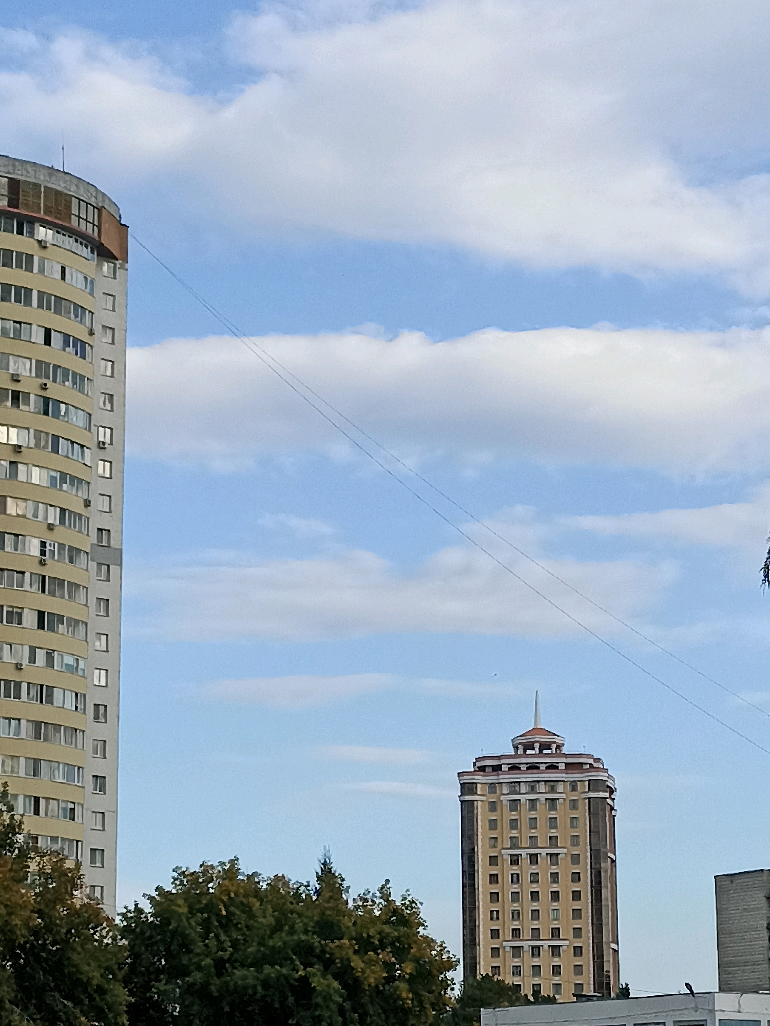 The image depicts an outdoor urban scene with a clear sky and scattered clouds. In the foreground, on the left, is the side of a tall, curved residential building with numerous windows. To the right and slightly further back is a distinct, taller, rectangular building with a decorative top and many windows. In the lower part of the image, a dense line of green trees spans across the width. The scene appears to be set in a city, likely Novosibirsk, Russia, given the architectural style of the buildings. There are no people visible in the image, and the focus is on the structures and natural elements. Two parallel wires are visible stretched across the sky, diagonally from the upper left to the lower right of the frame.