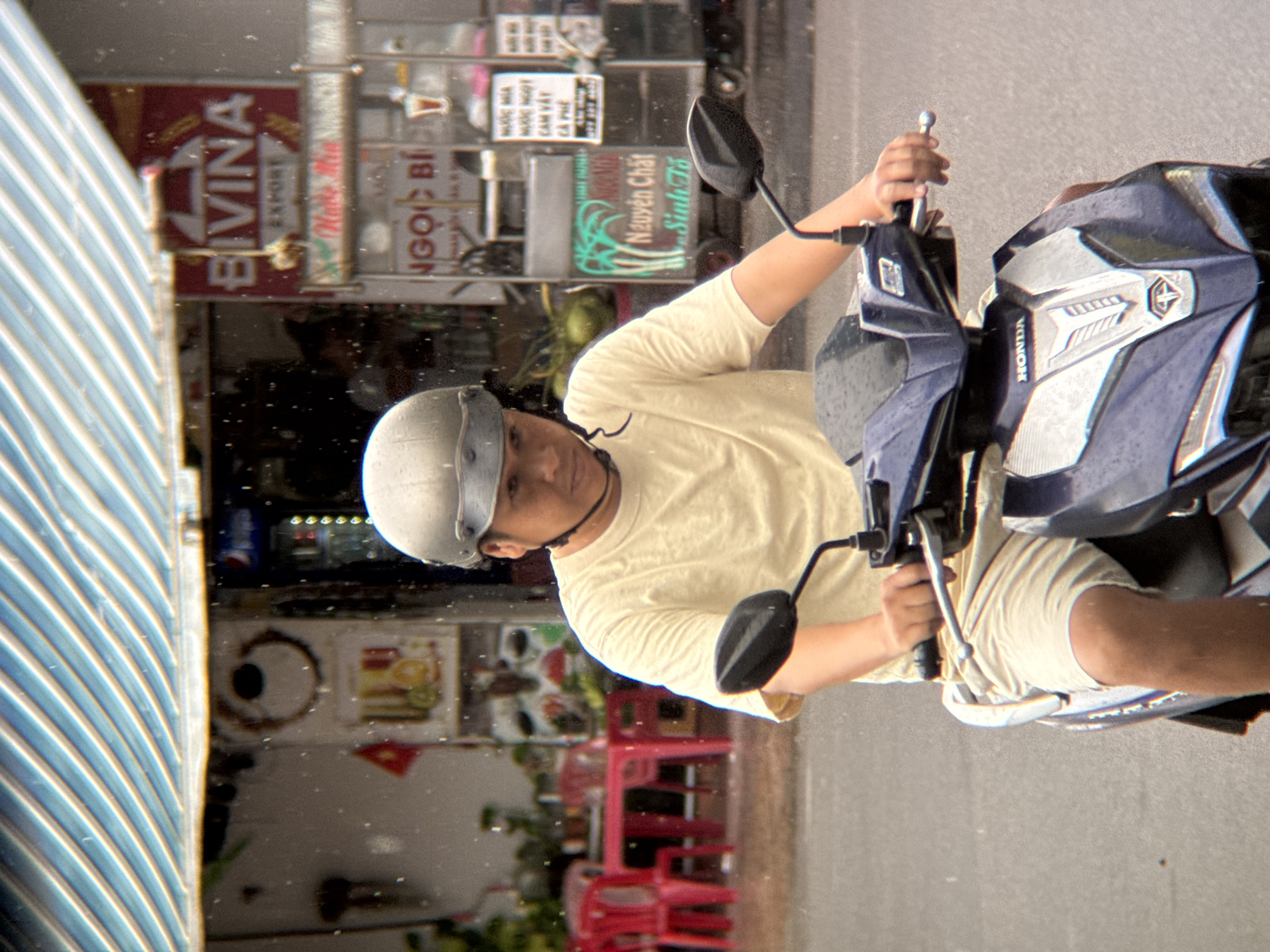 In Nha Trang, Vietnam, a person wearing a grey helmet and a light yellow t-shirt rides a blue and silver Honda motorcycle on a wet street, indicated by water droplets on the surface and in the air. The person wears light-colored shorts. In the background, red tables and chairs suggest an outdoor seating area for a cafe or restaurant. Above the street scene, a striped awning, predominantly blue and white, is visible. Further in the background, there are signs with Vietnamese text, including one that reads "BIVINA EXPORT" and another with "NGUYEN CHAT SINH TO," indicating food and drink vendors. The overall atmosphere suggests it is raining, and the city is bustling with activity.