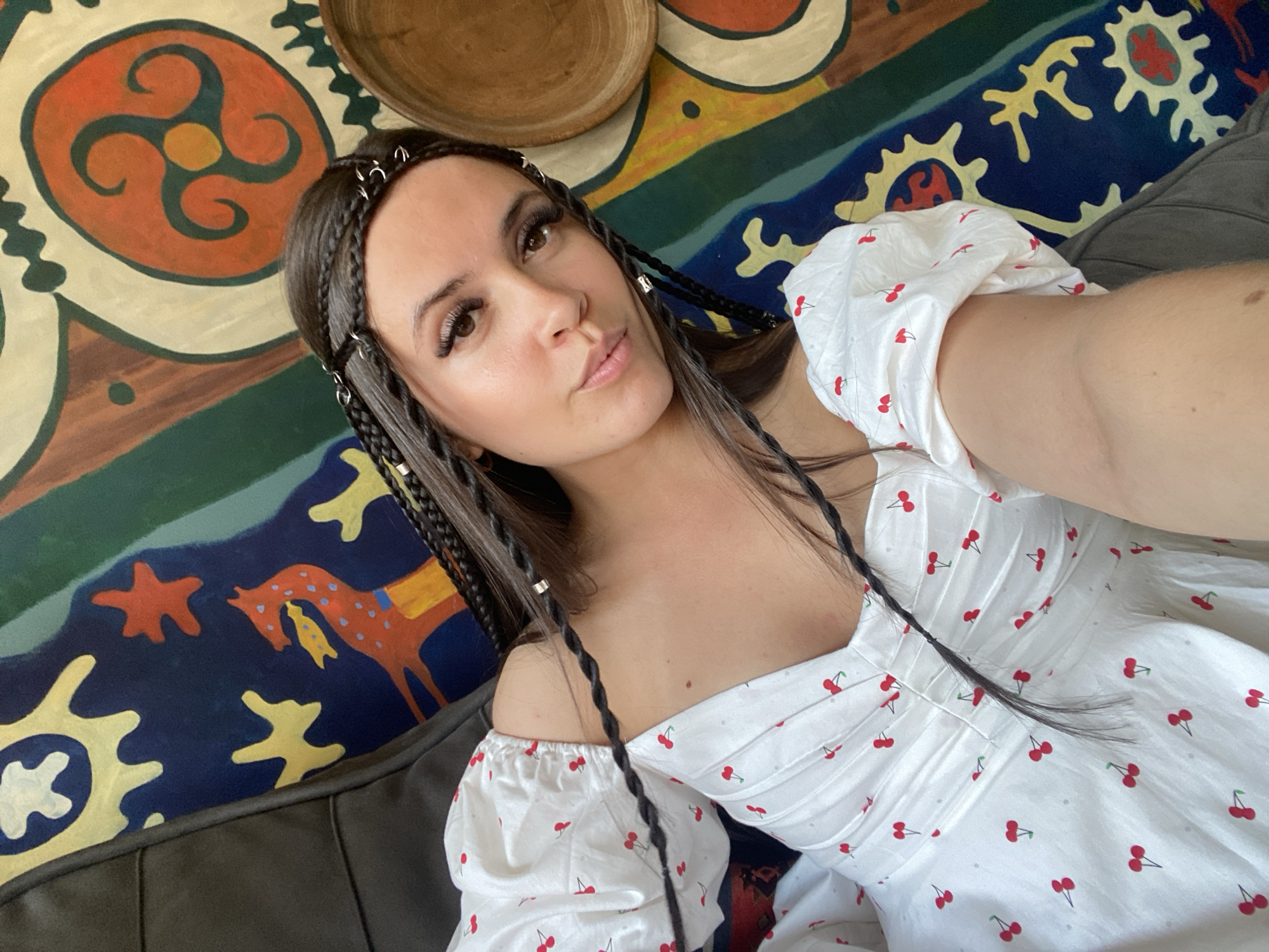 A young woman with dark hair styled in braids adorned with silver rings takes a selfie while sitting on a couch. She is wearing a white dress with a cherry print. The background features a colorful, patterned tapestry and a wooden bowl. The scene appears to be indoors, possibly in Makhachkala, Russia, given the cultural motifs in the tapestry which may be inspired by local or Central Asian designs.