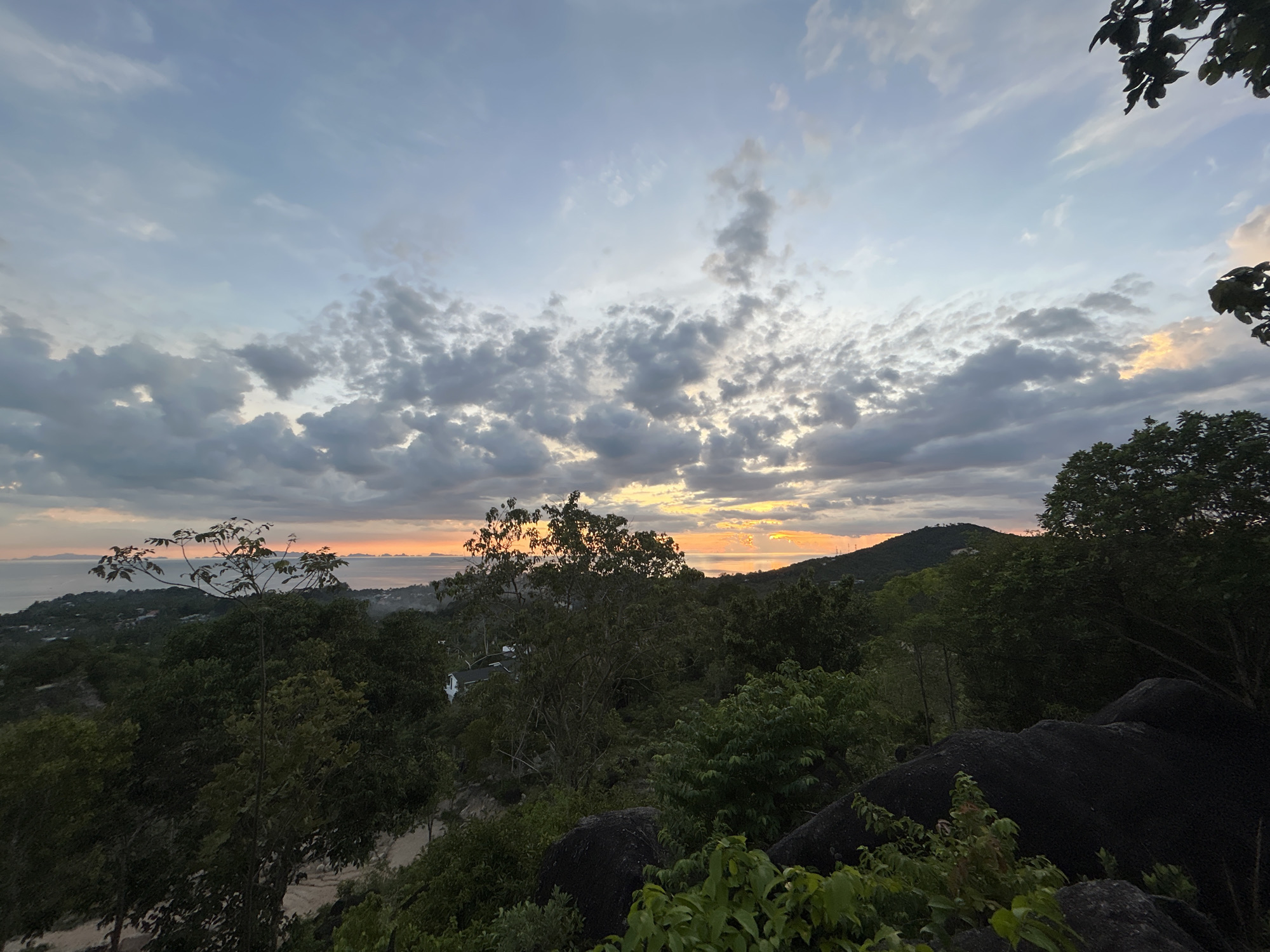 This is a wide-angle, eye-level shot of a tropical landscape at sunset, viewed from an elevated position overlooking the sea. The sky is filled with dramatic clouds, ranging from light grey to darker hues, with streaks of orange and yellow light breaking through on the horizon, indicating the setting sun. The foreground is dominated by lush green foliage and large, dark grey boulders. In the mid-ground, a dense forest covers rolling hills, with scattered glimpses of buildings and infrastructure, suggesting a populated area. The distant background features a calm body of water, likely the Gulf of Thailand, with a chain of islands visible on the horizon. The overall impression is one of natural beauty and a serene, possibly remote, tropical setting. The location is identified as Tambon Ko Pha Ngan, Thailand, known for its scenic beaches, lush jungles, and mountainous terrain.