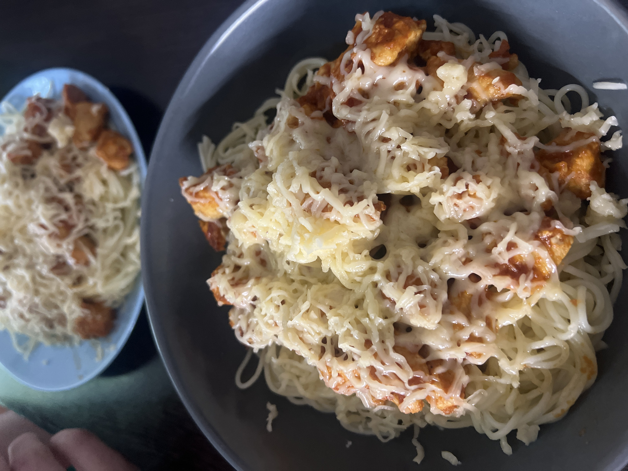 The image shows two bowls of spaghetti with melted cheese and fried chicken. The larger bowl is in the foreground and is a grey color. The smaller bowl is behind and to the left, and is light blue. A small portion of a finger is visible in the bottom left corner. The background is dark and out of focus. There is no evidence to suggest that this image was taken in Krasnoyarsk, Russia.