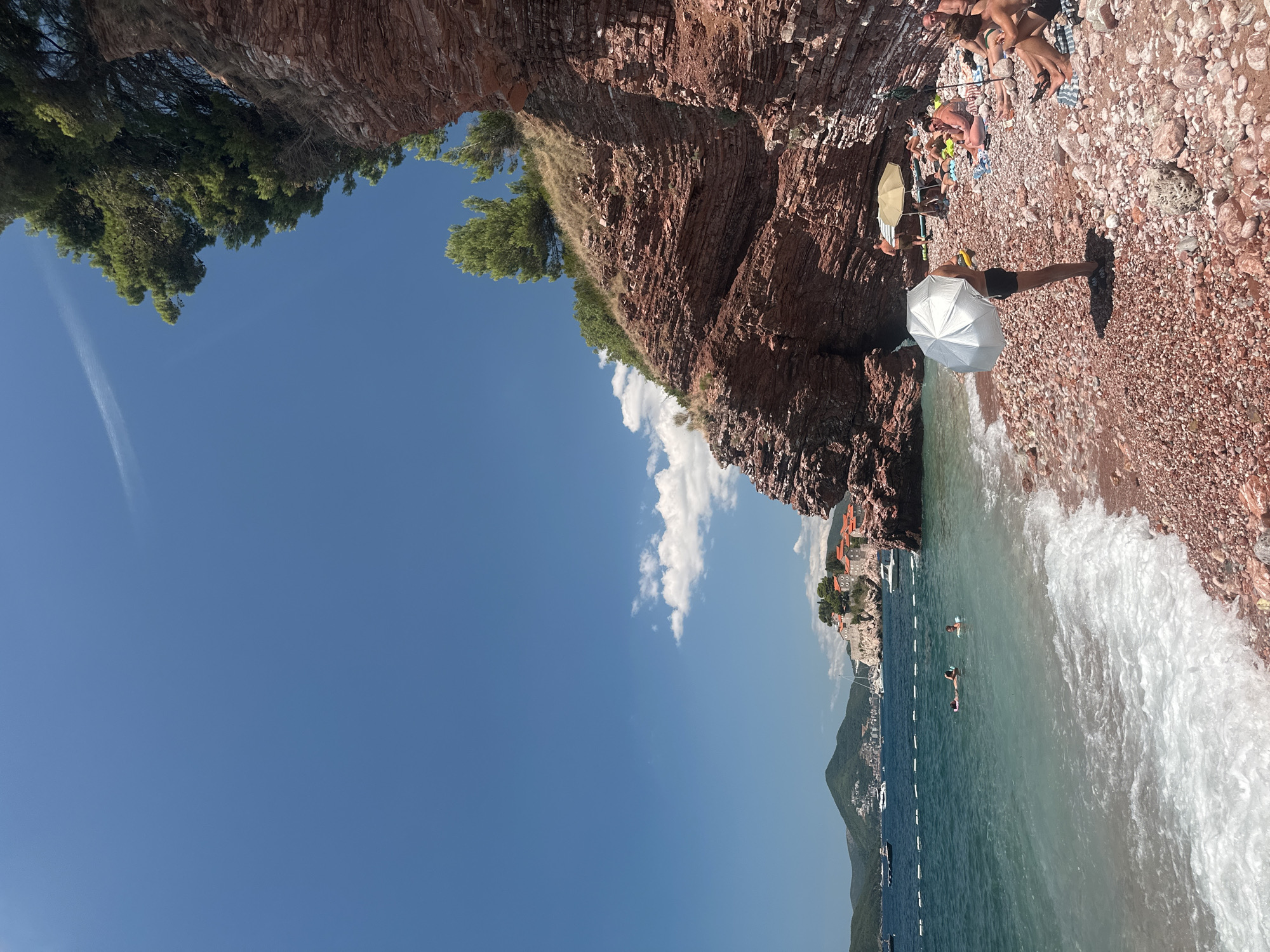 This is a wide shot of a pebbly beach on a sunny day in Sveti Stefan, Montenegro. The beach is situated at the base of a steep, rocky cliff with layered red rock formations and sparse green vegetation. The Adriatic Sea is a clear turquoise, with gentle waves breaking on the shore. In the distance, several boats are anchored, and a small town with orange-roofed buildings is visible on a nearby island connected to the mainland by a causeway.
On the beach, several people are relaxing. Some are lying on towels or sitting under umbrellas, while others are in the water. One person in the foreground stands holding a silver umbrella. The overall atmosphere is one of a relaxed summer vacation.