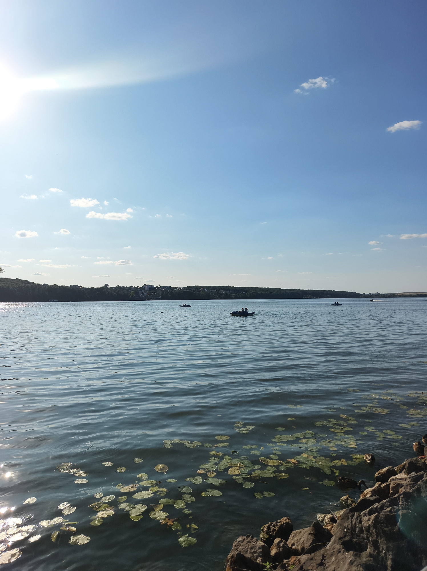 The image depicts a serene lake scene on a bright, sunny day. The sky is a clear blue with a few wispy white clouds, and the sun is shining, casting a bright glare on the water. In the foreground, the rocky shore of the lake is visible, with several water lilies scattered on the surface of the water. Further out on the lake, there are several small boats with people in them. The far shore of the lake is lined with trees and some buildings, suggesting a populated area. The overall atmosphere is one of tranquility and natural beauty. The scene is likely located at the Ternopil Reservoir in Ternopil, Ukraine, a popular recreational area.