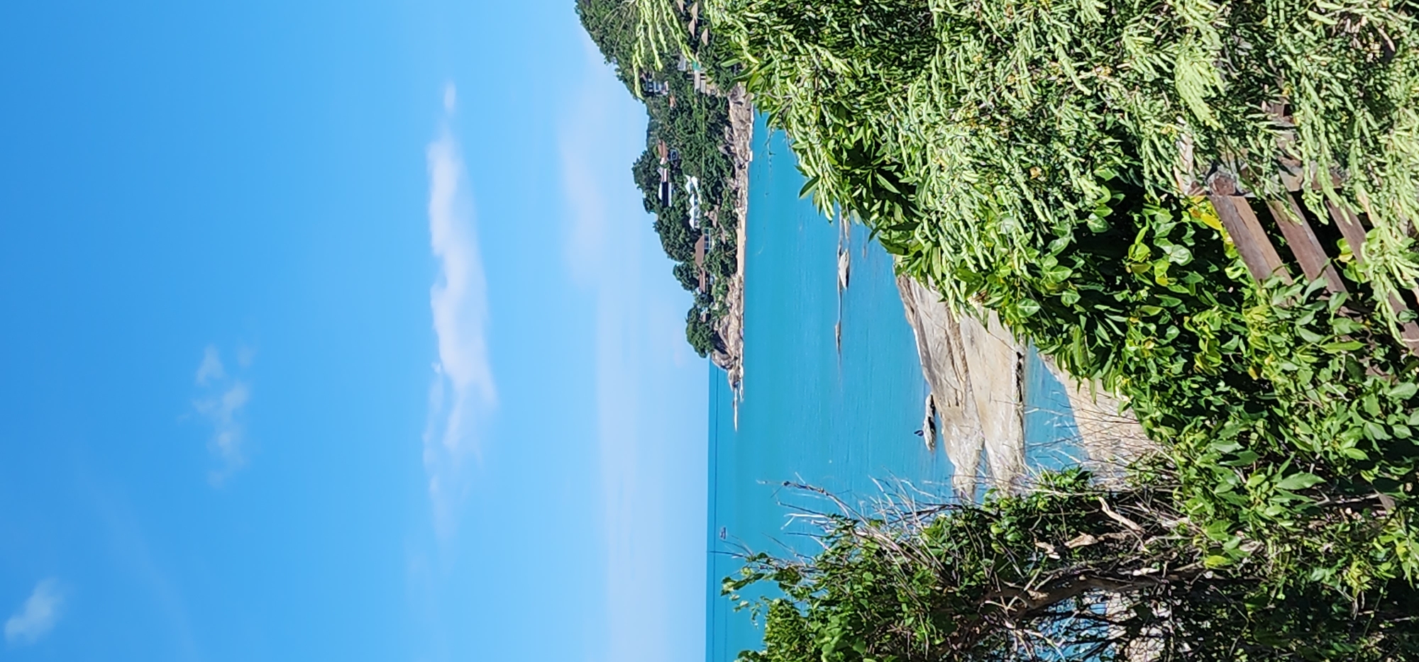 The image depicts a coastal scene in Baan Chaweng Noi, Thailand. The foreground is dominated by lush green foliage and a wooden railing, obscuring the lower portion of the frame. Beyond the vegetation, a body of clear, turquoise water stretches towards a rocky coastline. Several large, flat rocks are visible at the water's edge, some partially submerged. On one of these rocks, a solitary dark figure is seated. The middle ground features a headland covered in dense green trees and dotted with buildings, indicative of a residential area or resort. The distant horizon is marked by the meeting of the bright blue sky and the sea, with a few wispy white clouds present. The overall impression is one of a serene tropical landscape.