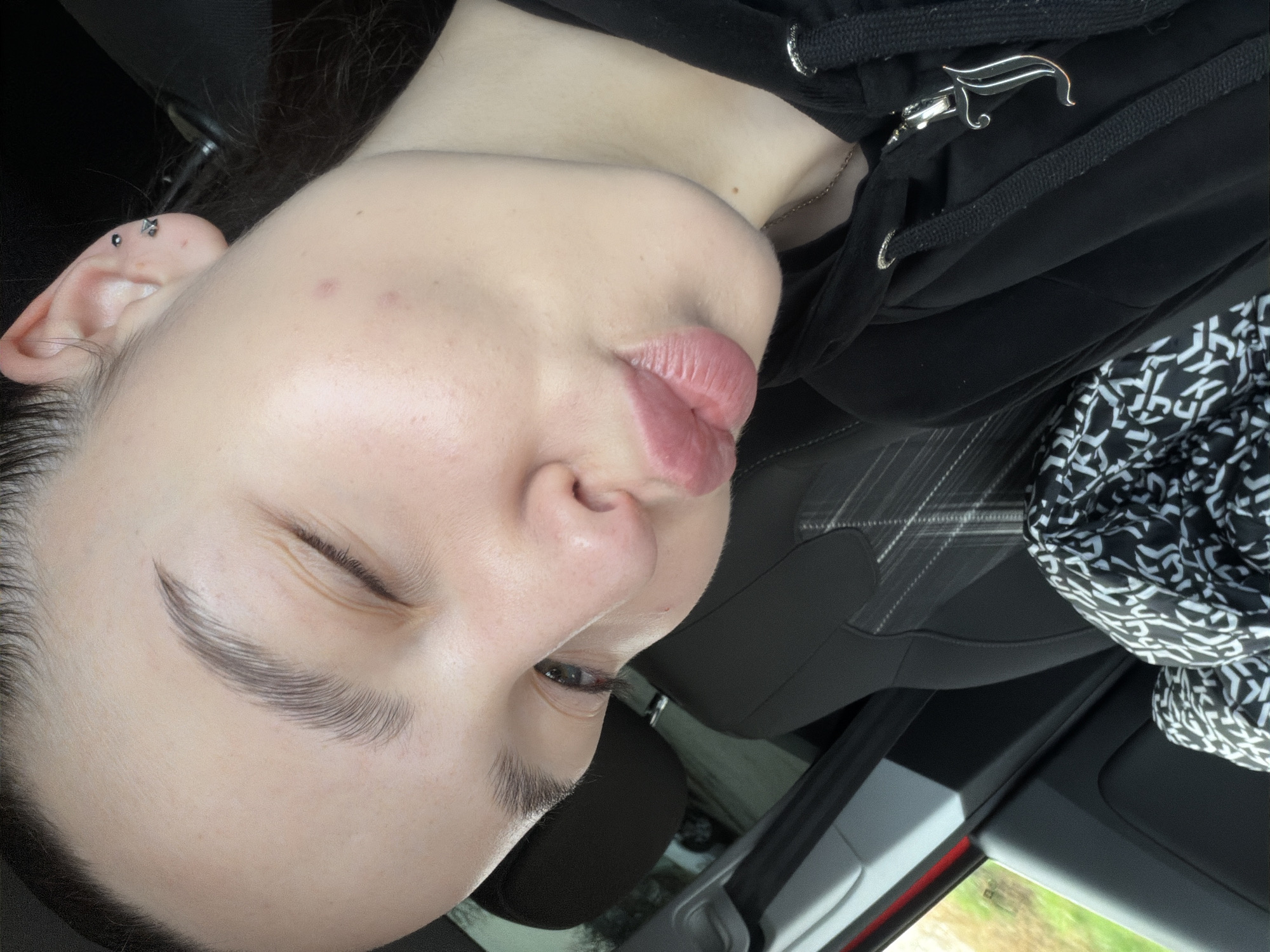 The image depicts a person with fair skin, likely in a car. They are wearing a black hooded sweatshirt with a silver "J" zipper pull. A black and white patterned scarf is visible near the seat. The person has an eyebrow piercing and an ear piercing. The location is inside a car, with glimpses of an exterior view of Busmantsi, Bulgaria, visible through the window.