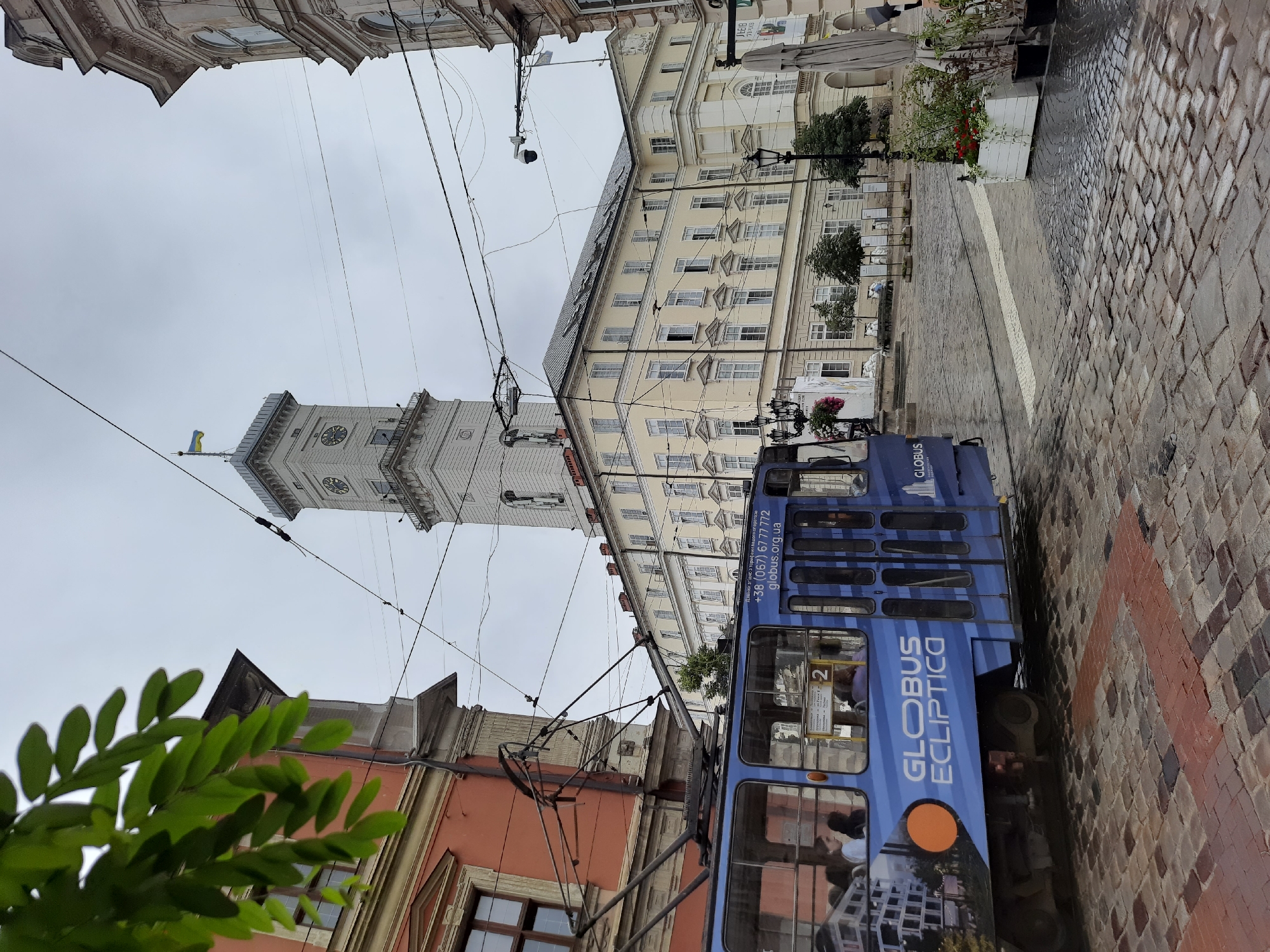 The image captures a street scene in Lviv, Ukraine, with a blue tram prominently featured in the foreground. The tram has the "GLOBUS ECLIPTICO" logo and a phone number visible on its side. In the background, a tall, light-colored clock tower with the Ukrainian flag on top stands against a cloudy sky. Adjacent to the tower is a light yellow building with many windows. The street is paved with cobblestones, and overhead, a network of power lines and tram wires crisscrosses the sky. Several people are visible inside the tram, and in the distance, other buildings and street elements like lampposts, trees, and planters can be seen. The overall atmosphere suggests an urban environment on a possibly overcast day.