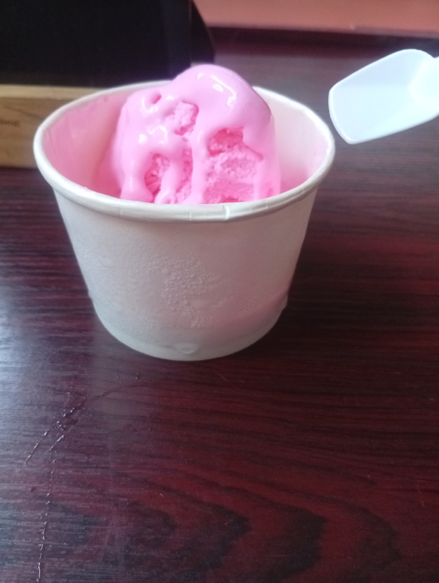 A white paper cup filled with vibrant pink ice cream sits on a dark, reddish-brown wooden table. The ice cream appears slightly melted, with glossy pink liquid visibly dripping down its textured surface. The exterior of the paper cup shows numerous small condensation droplets, indicating its cold temperature. To the right of the cup, a small, plain white plastic spoon is visible. The wooden table features a distinct grain pattern, and subtle, dried liquid streaks can be seen on its surface, particularly to the left of the cup. In the blurred background on the left, a dark, indistinct object, possibly a piece of furniture or a box, is partially visible. No individuals are present in the image. The setting is an indoor environment, likely in Howayi, Nigeria, with soft lighting illuminating the dessert. No legible text is visible.