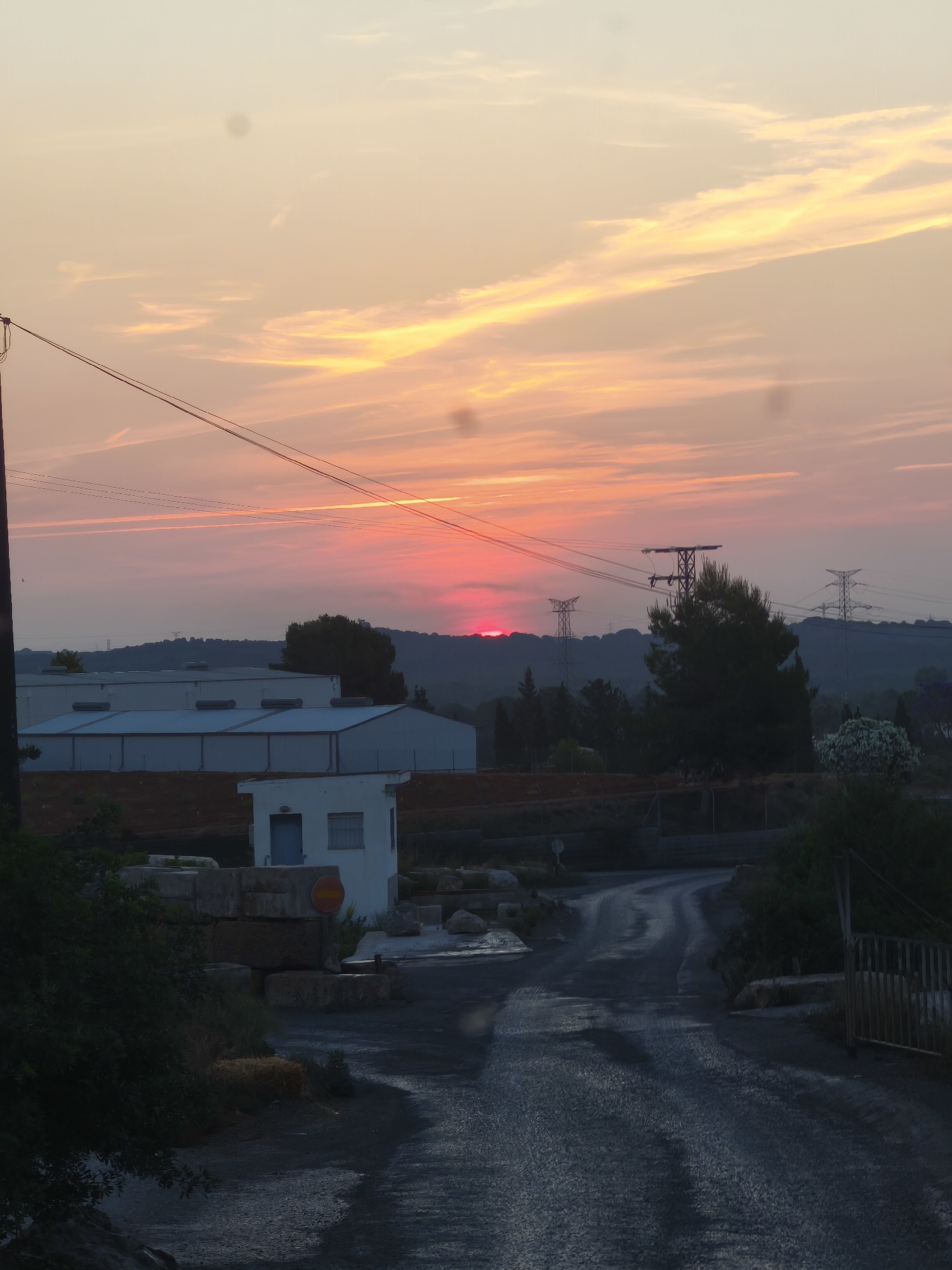 The image depicts a street scene at dusk in Riba-roja de Túria, Spain. The sun is setting, casting a red and orange glow across the sky. In the background, there are low hills, industrial buildings with flat roofs, and electrical pylons. A small white building is visible on the left side of the road, along with a stack of stones. The road itself is asphalt and appears wet, leading away from the camera. Vegetation lines the sides of the road, including shrubs and trees. A metal gate is visible on the right side of the road. Power lines stretch across the sky. No people are visible in the image.