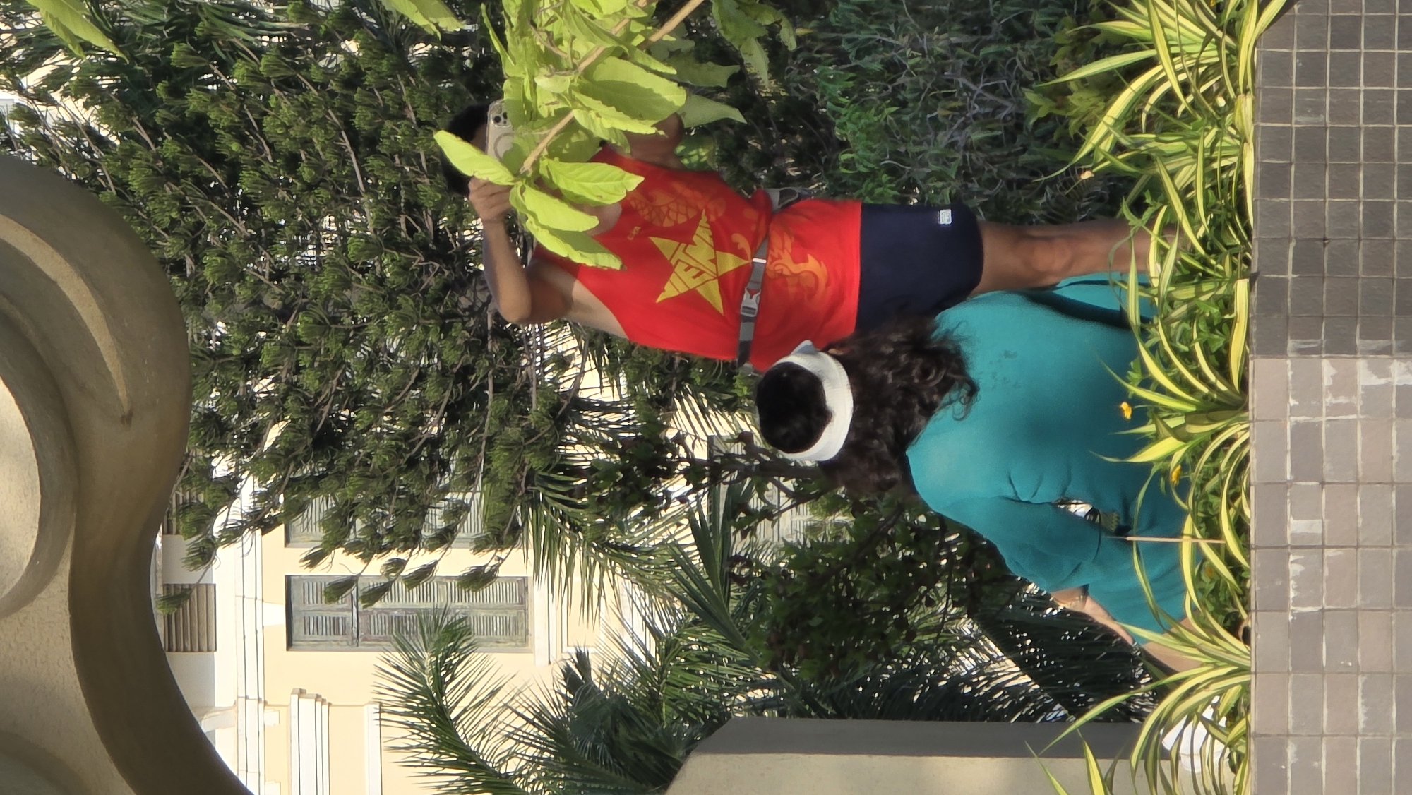The image depicts an outdoor scene featuring two individuals amidst lush greenery and architecture, oriented horizontally for viewing. On the left, a man is captured mid-action, holding a silver smartphone up to take a picture or video. He wears a vibrant red sleeveless top prominently featuring a large yellow star, reminiscent of the Vietnamese flag, along with gold or orange dragon-like patterns. He also has on dark shorts and a white headband. His skin appears tanned, suggesting outdoor exposure.
To the right, a woman with dark, curly hair, also wearing a white headband, is seen from behind. She has on a teal or turquoise long-sleeved top and appears to be crouching or bending forward, possibly examining something on the ground or within the foliage.
The setting is characterized by abundant green vegetation, including tall conifer-like trees, palm fronds, and spiky green and yellow plants bordering a gray tiled area on the right. In the background, light-colored buildings with louvered windows are visible, suggesting an urban or semi-urban environment. A large, curved, light-beige concrete or stone structure partially frames the left side of the image. The bright, even lighting indicates a clear, sunny day, likely midday or afternoon. The man's shirt, bearing the yellow star on red, strongly suggests the location is Vietnam, consistent with the provided context of Nha Trang. No explicit legible text is visible other than the national emblem design on the man's shirt.