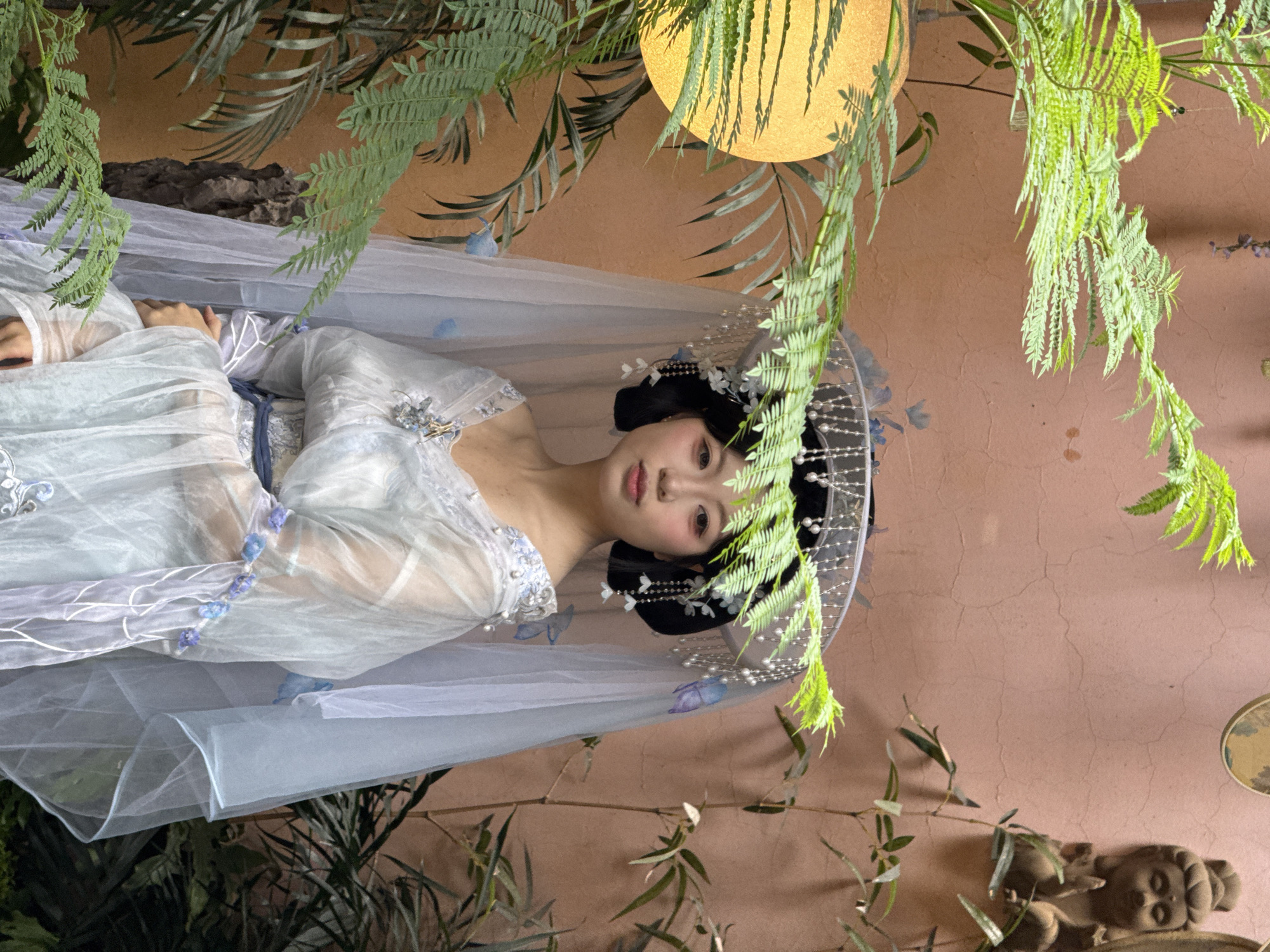 The image shows a woman in a traditional Chinese dress lying down outdoors. The location is likely in a garden setting, possibly in the Yuzhong District, China, given the style of the dress and the possible architectural details in the background. The woman is wearing a light-colored dress with intricate embroidery and floral embellishments. She has dark hair styled in a traditional updo, adorned with floral hair accessories. A sheer, veil-like fabric drapes around her. Ferns and other greenery are present in the foreground and background, creating a natural, possibly staged, environment. The background shows a textured, earth-toned wall, potentially a part of traditional Chinese architecture. A round, possibly decorative, light fixture can be seen above. There is also a small sculpture in the lower right corner of the image. The overall scene has a serene and artistic quality.