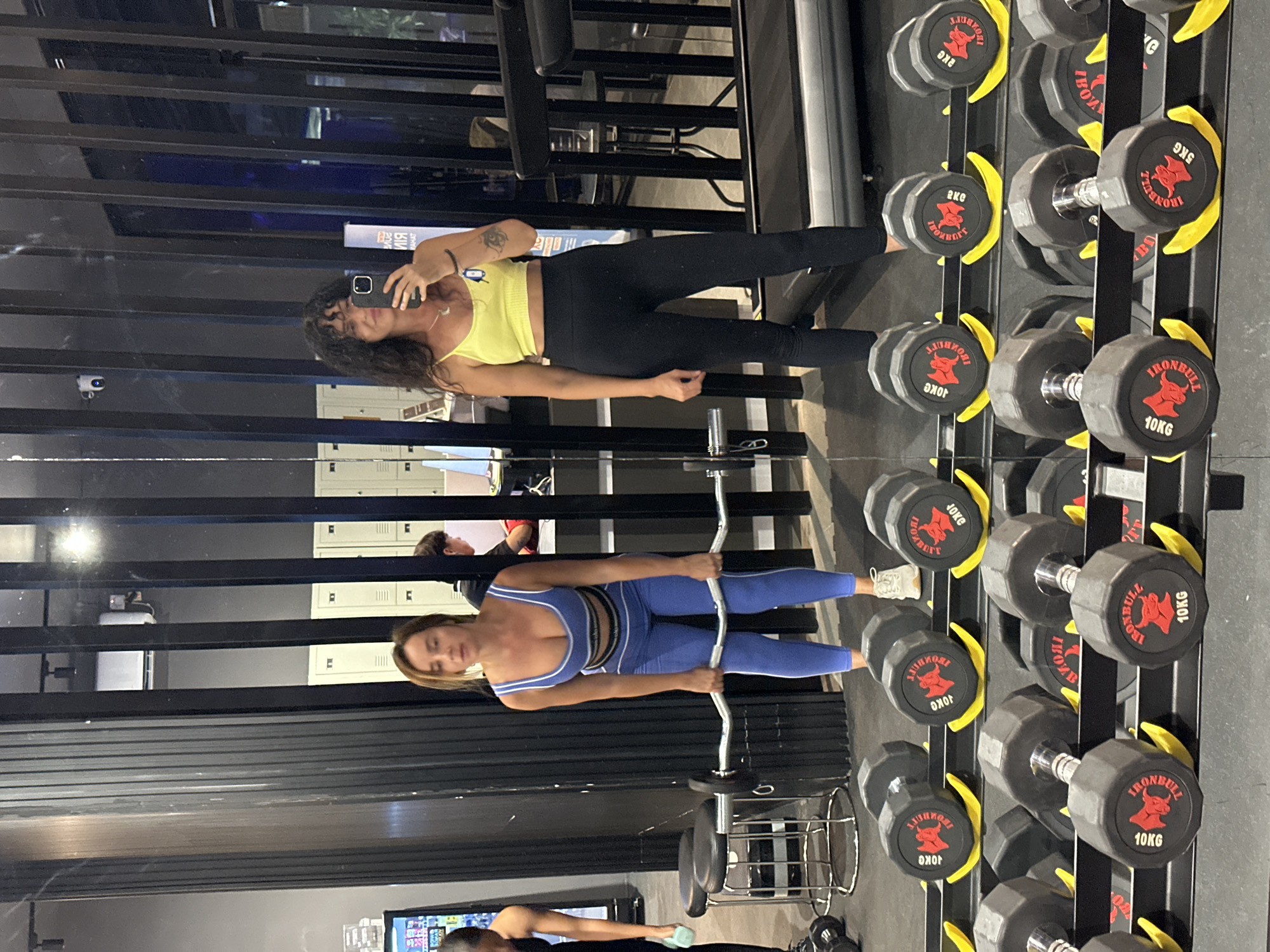 Two women are in a gym in Kuta Selatan, Indonesia. The woman on the left is wearing a blue athletic set and is holding a barbell with both hands. The woman on the right is wearing a yellow crop top and black leggings, and is taking a selfie with her phone. Dumbbells are displayed on a rack in the foreground. Lockers and a treadmill are visible in the background.