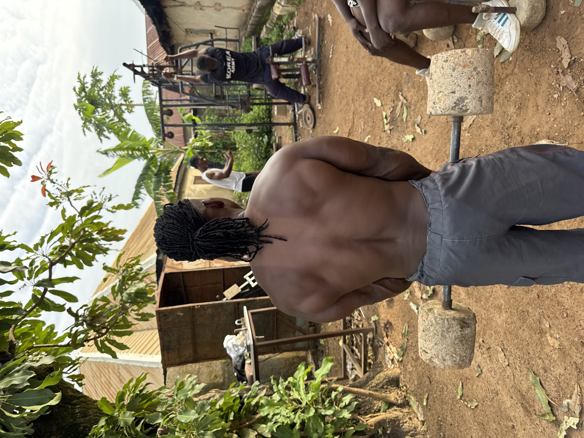 The image depicts an outdoor gym scene in the city of Akwanga, Nigeria. In the foreground, a muscular man with braided hair is seen from behind, holding a barbell loaded with two large, rough-hewn stone weights. He is wearing grey cargo pants. The ground is made of reddish-brown dirt, with scattered leaves. Behind him, two other men are visible. One man, wearing a black t-shirt with "KOREA ARMY" printed on it, is seated on a weightlifting machine, with his arms raised. Another man, wearing a white tank top, is standing near a metal structure, appearing to be performing an exercise. The gym equipment is rudimentary, made of metal pipes and concrete weights. In the background, there are buildings with corrugated metal roofs and trees, suggesting a semi-rural or residential setting. A table with some items on it is visible to the left of the foreground subject. The sky is overcast with some clouds.