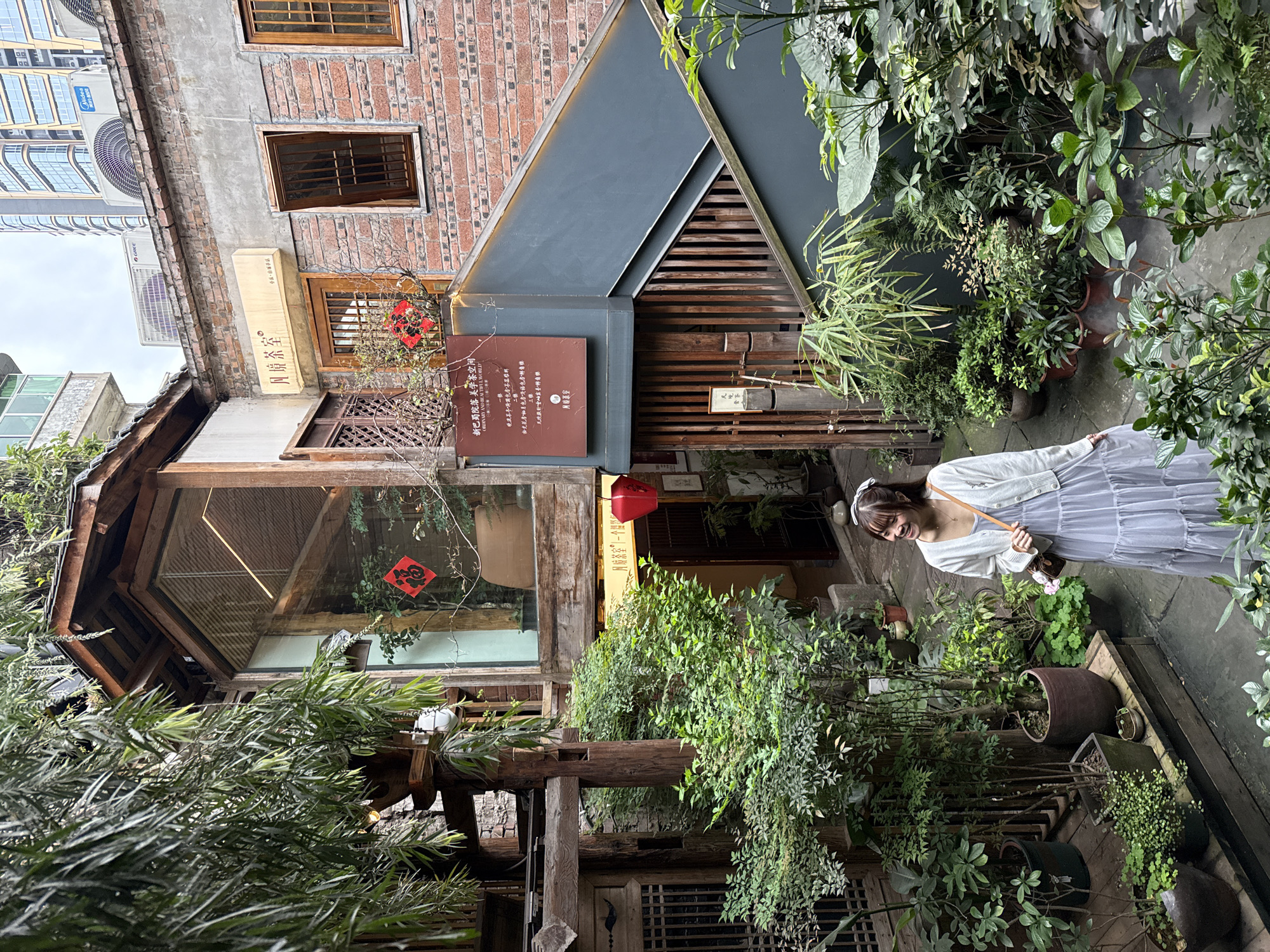 This image captures a lush urban courtyard in Yuzhong District, China, characterized by a blend of traditional architecture and modern elements under an overcast sky.
**Environment:** The central area is a densely vegetated courtyard featuring numerous potted plants of various species and sizes, arranged on stone paving and elevated wooden platforms. Some foliage hangs from overhead structures, and a fine mist emanates from ground-level plants on the right.
**Structures:**
* To the left and center, a multi-level wooden structure features exposed beams, large glass windows, and a sloped roof. A red square paper decoration with the Chinese character "福" (fú, meaning good fortune) is visible through one of the glass panes.
* To the right, a two-story building constructed of weathered red brick stands prominently. It has multiple rectangular windows with wooden frames and vertical grates. Above these windows, a horizontal sign displays Chinese characters "凡境茶室" and the English phrase "ORDINARY AND BEAUTIFUL WORLD". Below, a dark brown sign lists services for "新巴院落美学空间" (Xinba Courtyard Aesthetic Space) in Chinese and English.
* An exterior dark grey staircase with wooden slatted railings descends from the brick building into the courtyard.
* Above the brick building, the sky is partially visible, showing modern high-rise buildings in the distance and several outdoor air conditioning units (GREE, Midea, AUX) on the rooftop.
**Objects:** A red traditional Chinese lantern hangs near an entrance under an overhang of the central wooden structure.
**Person:** A young adult female, of East Asian ethnicity, stands in the lower-right portion of the courtyard. She has shoulder-length brown hair, tied back with a white bow. She wears a light-colored, possibly lavender or light grey, tiered maxi dress, covered by a white button-up cardigan. She carries a small, patterned brown shoulder bag. Her head is turned to her left (viewer's right), and she is smiling, appearing to look towards an unseen point of interest outside the frame. Her posture suggests she is either pausing or in motion, navigating the courtyard.