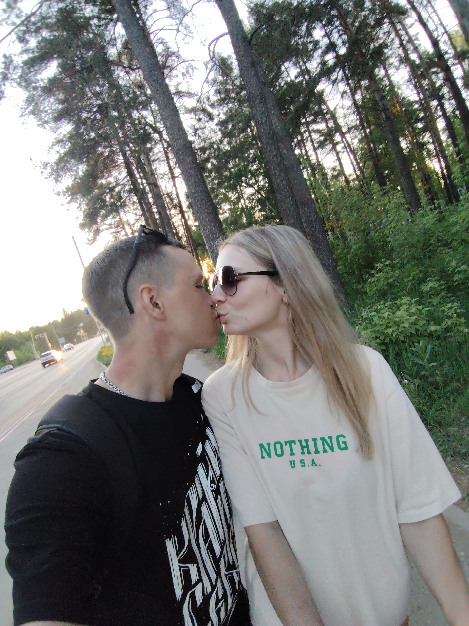 A couple is kissing on a sidewalk in Chaikovsky, Russia. The man wears a black t-shirt, a silver chain, and has sunglasses perched on his head. The woman wears sunglasses and a cream-colored t-shirt with the words "NOTHING U.S.A." printed in green. They are standing next to a road with cars visible in the distance. Behind them is a dense forest with tall trees. The lighting suggests it is late afternoon or early evening.