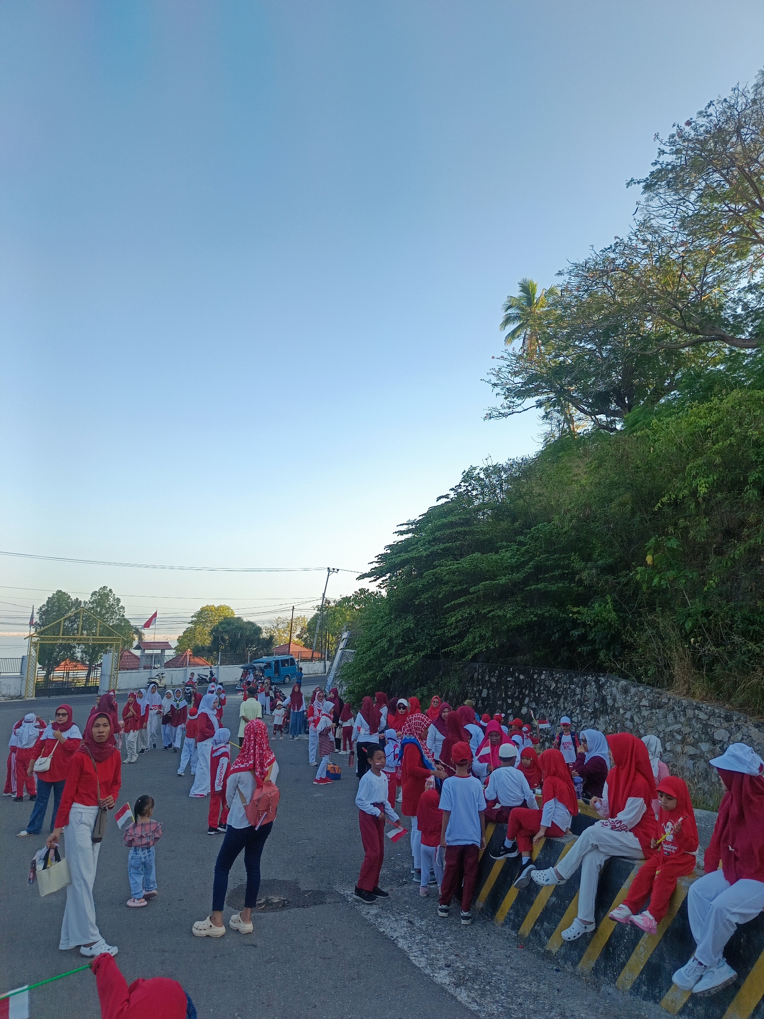 The image depicts a group of people, primarily children and adults, gathered outdoors in what appears to be a street or open area. Many are wearing red and white attire, which are the national colors of Indonesia, suggesting a patriotic event or celebration. Some individuals are holding small Indonesian flags. In the background, there are trees, a hill covered in vegetation, buildings, a blue van, and a body of water visible in the distance, hinting at a coastal or near-coastal location. The sky is clear and blue, suggesting it is daytime, likely morning or late afternoon due to the soft lighting. The presence of a yellow and black striped barrier indicates a roadside setting. The overall atmosphere seems festive and communal. The location is identified as Ilotidea, Indonesia.