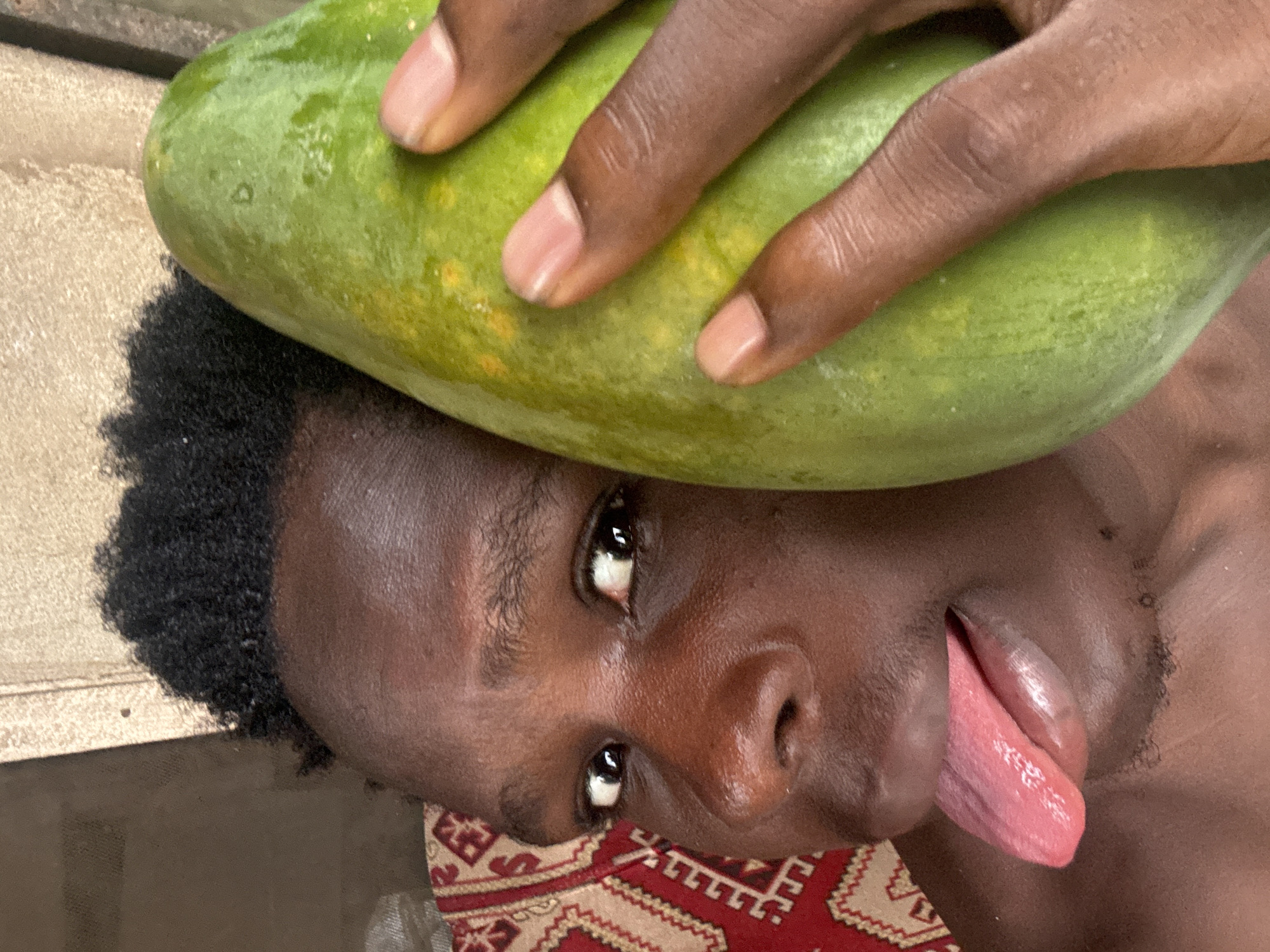 A close-up, angled photograph shows a person lying on a surface with a patterned fabric visible beneath their head. The person is holding a large, green papaya against their forehead with their hand. The person's tongue is extended. No specific location in Oboama, Nigeria, can be identified from the image.