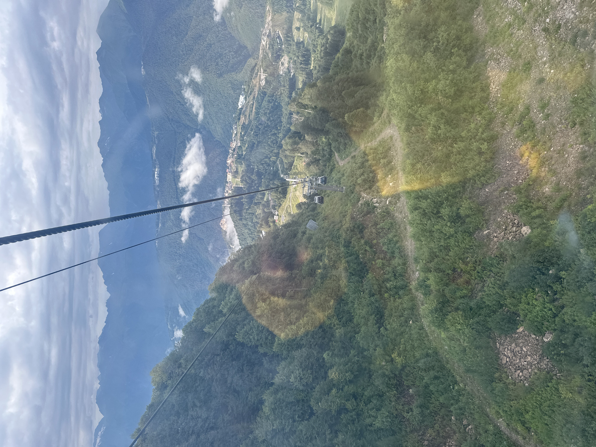 This image, taken from a cable car in Estosadok, Russia, offers a panoramic view of a mountainous landscape. The foreground is dominated by a steep, heavily wooded slope, characterized by lush green trees and scattered rocks. A winding dirt path is visible traversing the hillside. Further down, a cable car system is in operation, with several cabins suspended from a thick cable. In the mid-ground, the terrain opens up to reveal a valley dotted with buildings, suggesting a settlement or resort area. A series of ski lifts and infrastructure are also visible, hinting at recreational activities. The background is defined by a majestic range of mountains, their peaks partially obscured by clouds and haze. The sky is overcast, with clouds casting shadows on the landscape. The scene is imbued with a sense of natural beauty and human development integrated within a rugged alpine environment.
