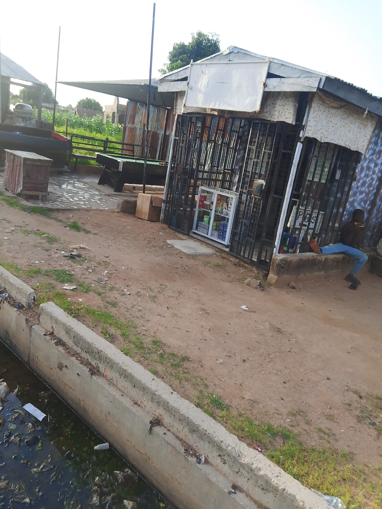 The image depicts an outdoor scene in Jalingo, Nigeria, likely a small marketplace or roadside establishment. The setting is characterized by a rough, sandy ground and a concrete ditch filled with murky water and debris running along the left side of the frame.
In the background, there are several structures with corrugated metal roofs and walls, suggesting a semi-permanent or improvised construction. One structure appears to be a small shop with a glass display case filled with various products, including what look like packaged goods and drinks. This shop is protected by metal bars, implying security measures.
In front of the shop, a person is seated on a low concrete ledge, wearing blue jeans and a dark top. They are looking towards the right side of the frame. Another person, partially visible, is also in the vicinity.
To the left of the shop, there is a pool table set up, indicating that leisure activities might be offered at this location. Nearby, a small wooden structure, possibly a storage box or bench, is visible.
The time of day appears to be late afternoon or early evening, given the soft lighting and long shadows. The weather seems to be clear. The overall atmosphere suggests a casual, everyday scene in a local Nigerian community. No legible text is visible in the image.