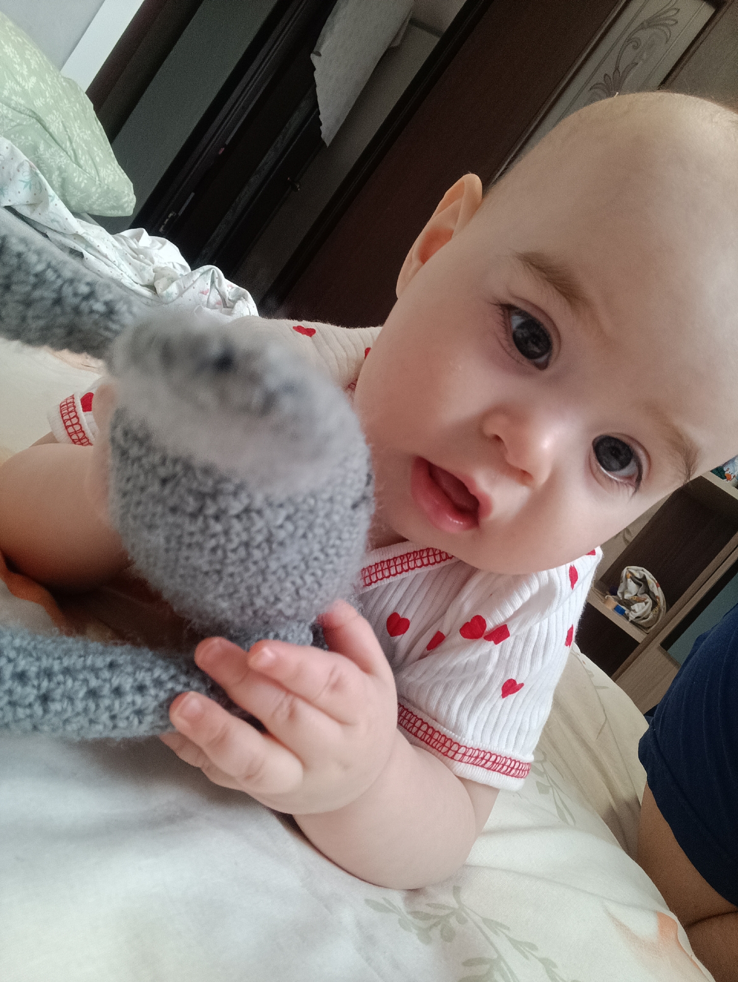 An infant with fair skin and dark, wide-set eyes lies on its stomach on a bed, engaging in what appears to be tummy time. The baby's head is tilted slightly upwards and to the right, looking directly at the camera with a curious and slightly surprised expression, its mouth slightly open. Its eyebrows are visible and well-defined. The baby wears a white, long-sleeved top adorned with numerous small red heart patterns and features red stitching along the cuffs.
The baby's small hands are wrapped around a grey, knitted or crocheted soft toy, which is partially out of focus and positioned in the foreground. The toy's texture is clearly visible, showing a consistent knitted stitch pattern. The bedding beneath the baby is light-colored, possibly white or cream, and appears soft. In the upper left background, a light green pillow with a faint pattern is visible, along with crumpled white fabric, likely more bedding.
The background setting suggests an indoor environment, possibly a bedroom in Noviy, Russia. Dark brown wooden furniture, resembling a wardrobe or shelves, is visible behind the baby, with a decorative light-colored panel above it. On one of the shelves to the right, a crumpled piece of light-colored cloth is visible. A dark blue fabric, possibly part of clothing worn by an unseen person, can be seen in the lower right corner. The lighting in the room appears bright and even, characteristic of daytime indoors. No text is visible in the image.