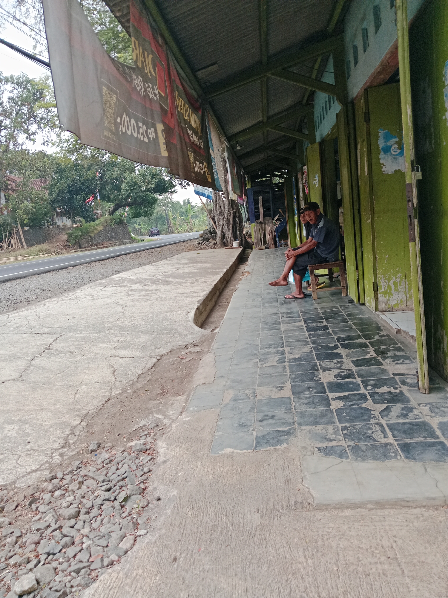The image captures a street scene in Bojongmengger, Indonesia, focusing on the facade of a row of shops. A man, wearing a dark collared shirt and shorts, sits on a wooden stool outside one of the shops, his feet bare and resting on the tiled walkway. He appears to be looking towards the viewer with an open-mouthed expression. Behind him, another person is partially visible. The shops have weathered green doors and a covered walkway with a corrugated metal roof, supported by green wooden beams. Banners hang from the roof, one prominently displaying text and what appears to be a QR code. The walkway is paved with dark square tiles, adjacent to a concrete sidewalk and a gravel-lined area that slopes towards a paved road. Trees and vegetation are visible beyond the road, with a dark-colored car driving in the distance. The overall scene suggests a casual, everyday setting in a rural or semi-rural area.