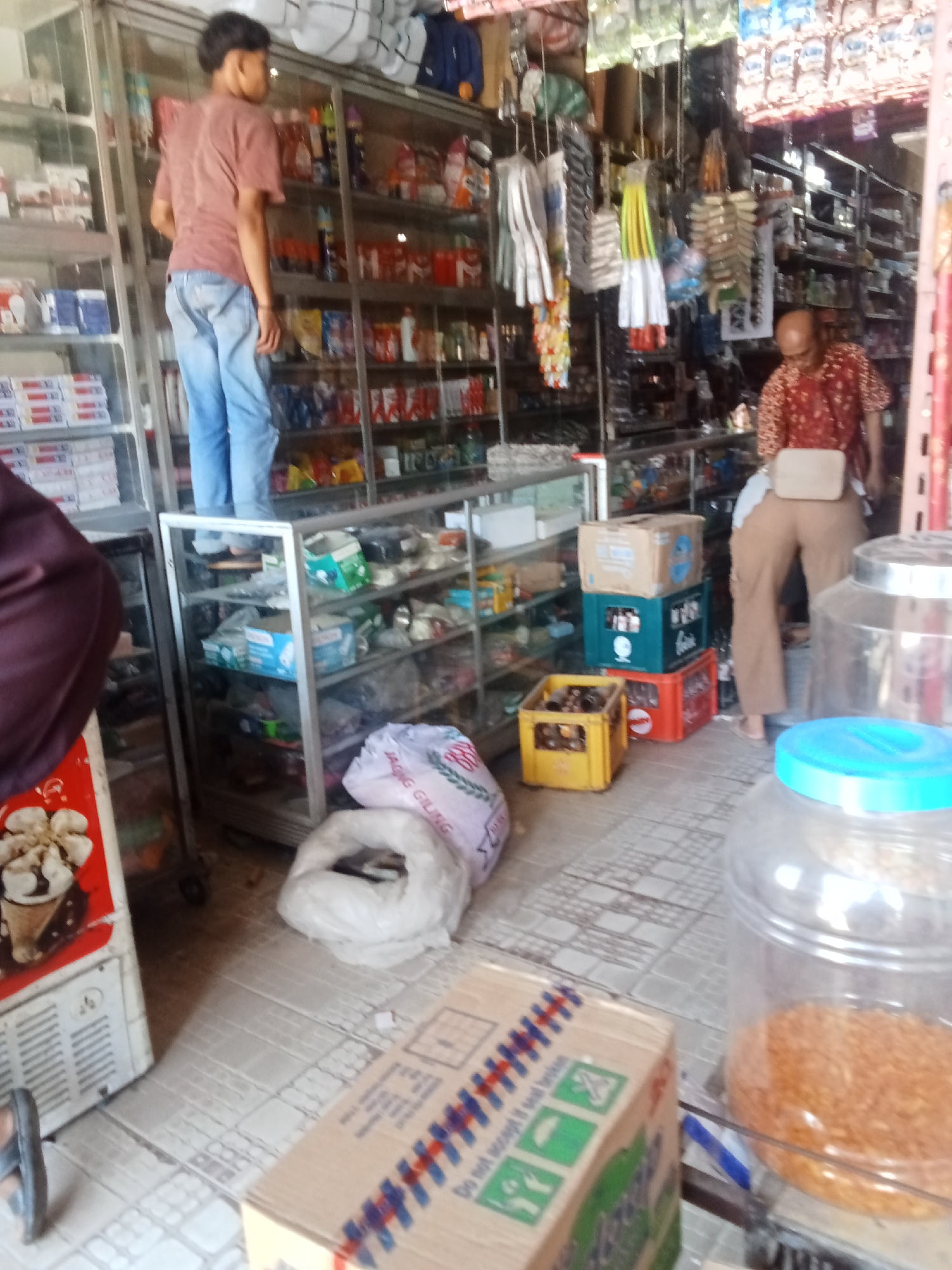The image captures the interior of a small convenience store, possibly a warung, located in Negeri Lama, Indonesia. The store appears to be organized with shelves stocked with various goods, including packaged food items, drinks, and household products. A young man, wearing a maroon t-shirt and blue jeans, is standing on a step stool, reaching for something on a high shelf. He is positioned in the central aisle of the store, facing away from the camera. To his right, a woman in a patterned red shirt and beige pants is seen from behind, appearing to be attending to customers or stocking shelves. She is wearing a beige fanny pack around her waist.
In the foreground, several items are placed on the floor. These include stacked crates of beverages, a yellow crate filled with bottles, and a large white plastic bag with text that is partially obscured but appears to read "JAGUNG GILING" (ground corn). A portion of a red and white freezer with an image of ice cream is visible on the left side of the frame. Further to the right, a large clear plastic jar filled with a granular orange substance, likely spices or food ingredients, is prominently displayed. The floor is tiled with a pattern of square tiles. The overall impression is that of a busy, functional retail space typical of a local Indonesian community.