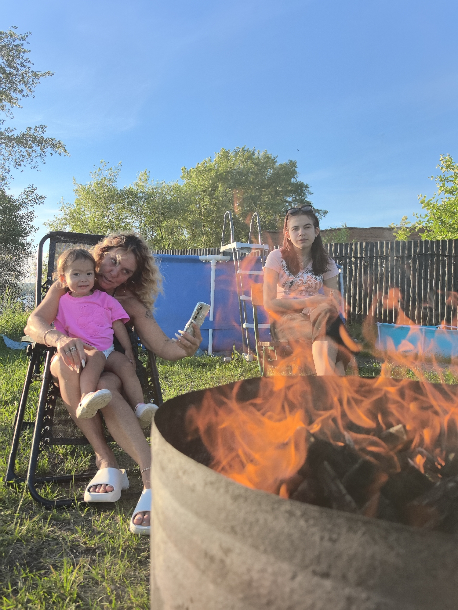 A woman with curly blonde hair is sitting in a black reclining chair outdoors, holding a young child on her lap. Both are looking at a cell phone held by the woman, who appears to be taking a selfie. The child is wearing a pink t-shirt. The woman is wearing a necklace and has painted fingernails. Her legs are crossed and she is wearing white slides. The child is wearing white sneakers. To the right of the chair, another woman with dark hair is sitting in a similar chair, looking towards the camera. She is wearing a pink t-shirt and sunglasses on her head. In the foreground, a metal fire pit with a large fire is visible, obscuring the bottom part of the scene. The background shows a blue swimming pool with a ladder, a wooden fence, and green trees. The sky is clear and blue. The scene appears to be a backyard in Chaikovsky, Russia, during daylight.