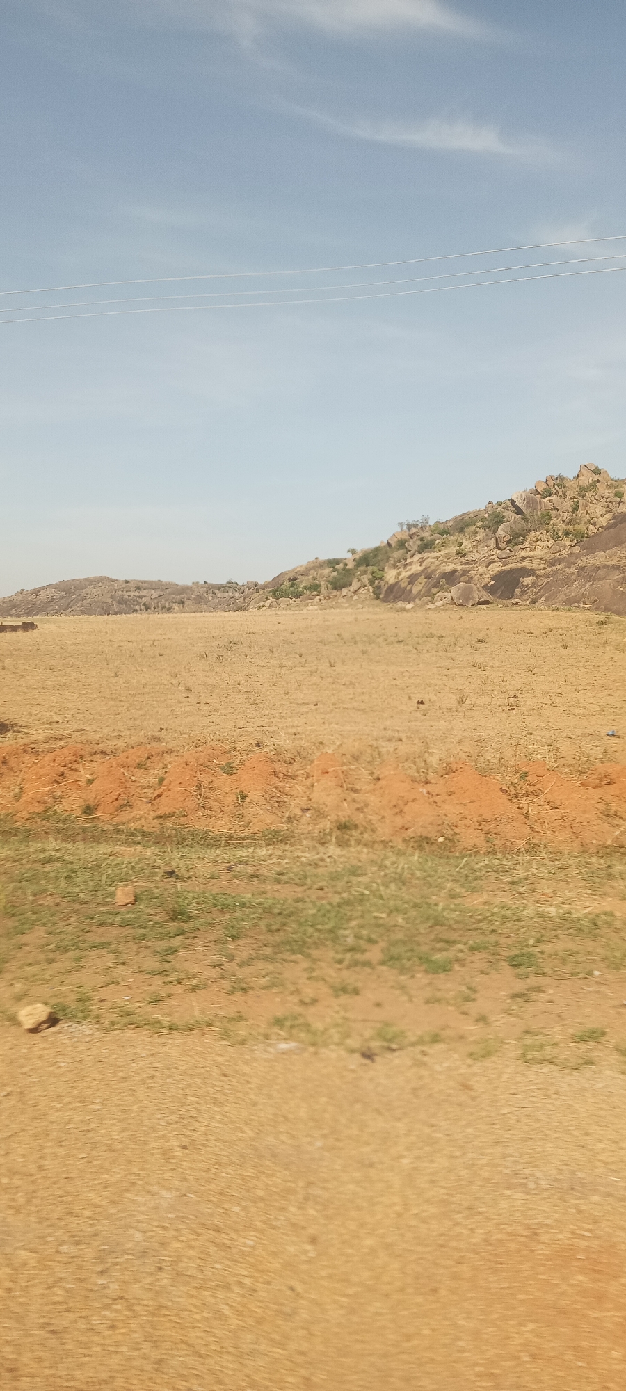 The image displays a broad outdoor landscape under a pale blue sky with faint, wispy clouds, suggesting clear daytime conditions. The foreground features dry, sandy soil interspersed with patches of sparse green grass. A prominent horizontal band of reddish-brown earth with small mounds, possibly termite hills or eroded soil, crosses the mid-foreground. Beyond this, a wide expanse of dry, yellowish-brown land extends towards the horizon, indicating an arid or dry environment.
In the background, a series of low, rocky hills and outcrops rise, covered with scattered grey and brown boulders and sparse green vegetation, including shrubs or small trees. Three thin, white power lines are visible stretching horizontally across the upper portion of the sky, hinting at nearby infrastructure. No human activity, people, or discernible objects other than the natural landscape and power lines are visible. The overall scene conveys a sense of rugged natural beauty and openness typical of a dry, sunny region, consistent with the specified location of Barakin Kuru III, Nigeria. There is no visible text.