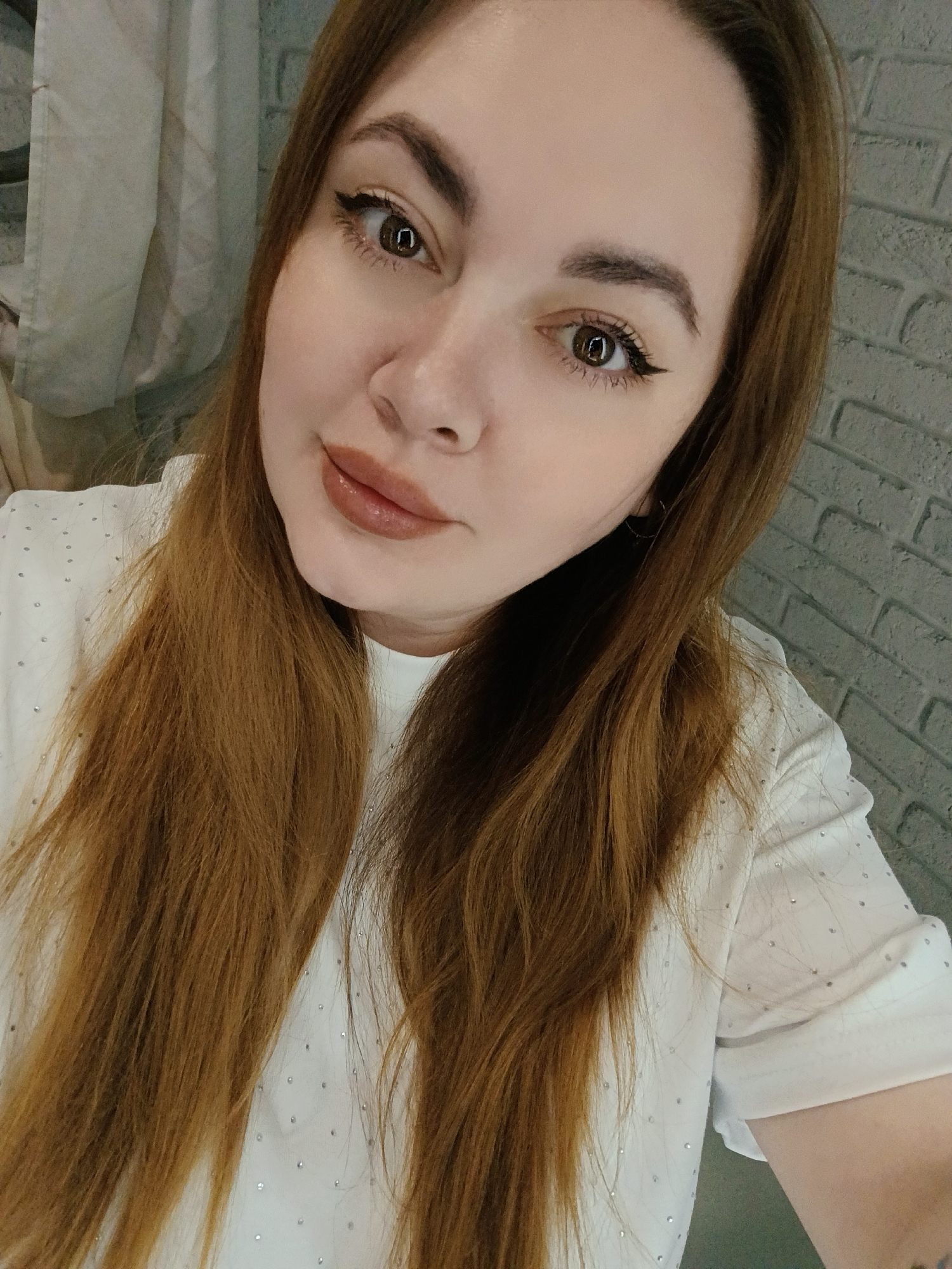 A selfie portrait shows a young woman with long, auburn hair. She has fair skin, brown eyes with black eyeliner, and wears a nude-toned lipstick. She is wearing a white blouse with small, scattered embellishments. The background features a textured, gray brick wall on the right and a white, draped fabric on the left. Based on previous data, the location is Chaikovsky, Russia.
