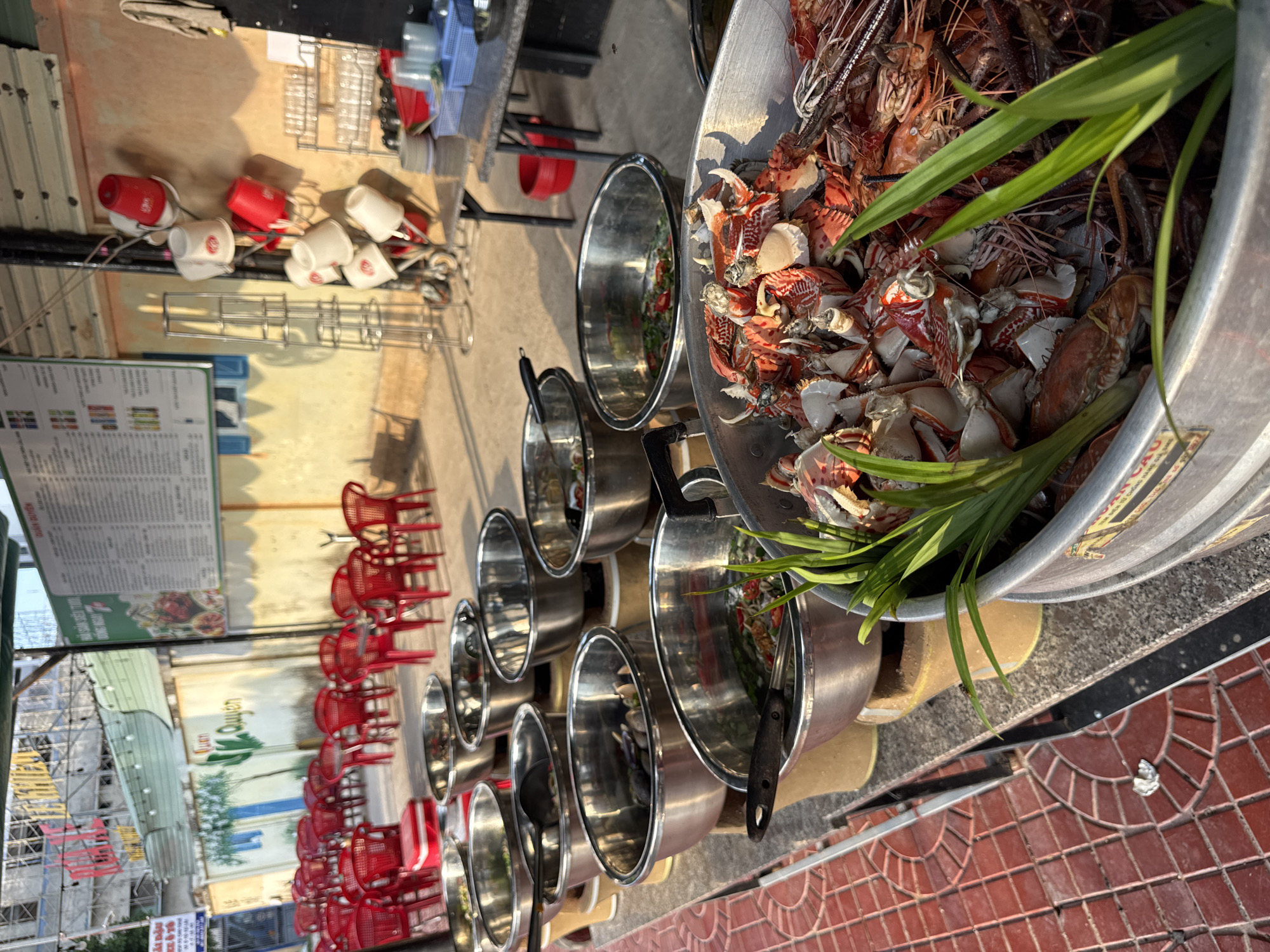 An outdoor food stall in South Quy Nhon Ward, Vietnam, displays a variety of seafood in large metal bowls. The stall is set up with rows of red plastic chairs and tables, suggesting it's a dining area. In the foreground, a large metal bowl is filled with cooked crabs and shrimp, garnished with green leaves. Behind it, several other bowls contain what appears to be seafood dishes in broth, with ladles resting inside. The ground is paved with red tiles. In the background, a menu board is visible on a wall, and a rack of white and red buckets hangs from a pole. The scene is illuminated by natural light, indicating it is daytime. There are no people immediately visible in the immediate vicinity of the food.