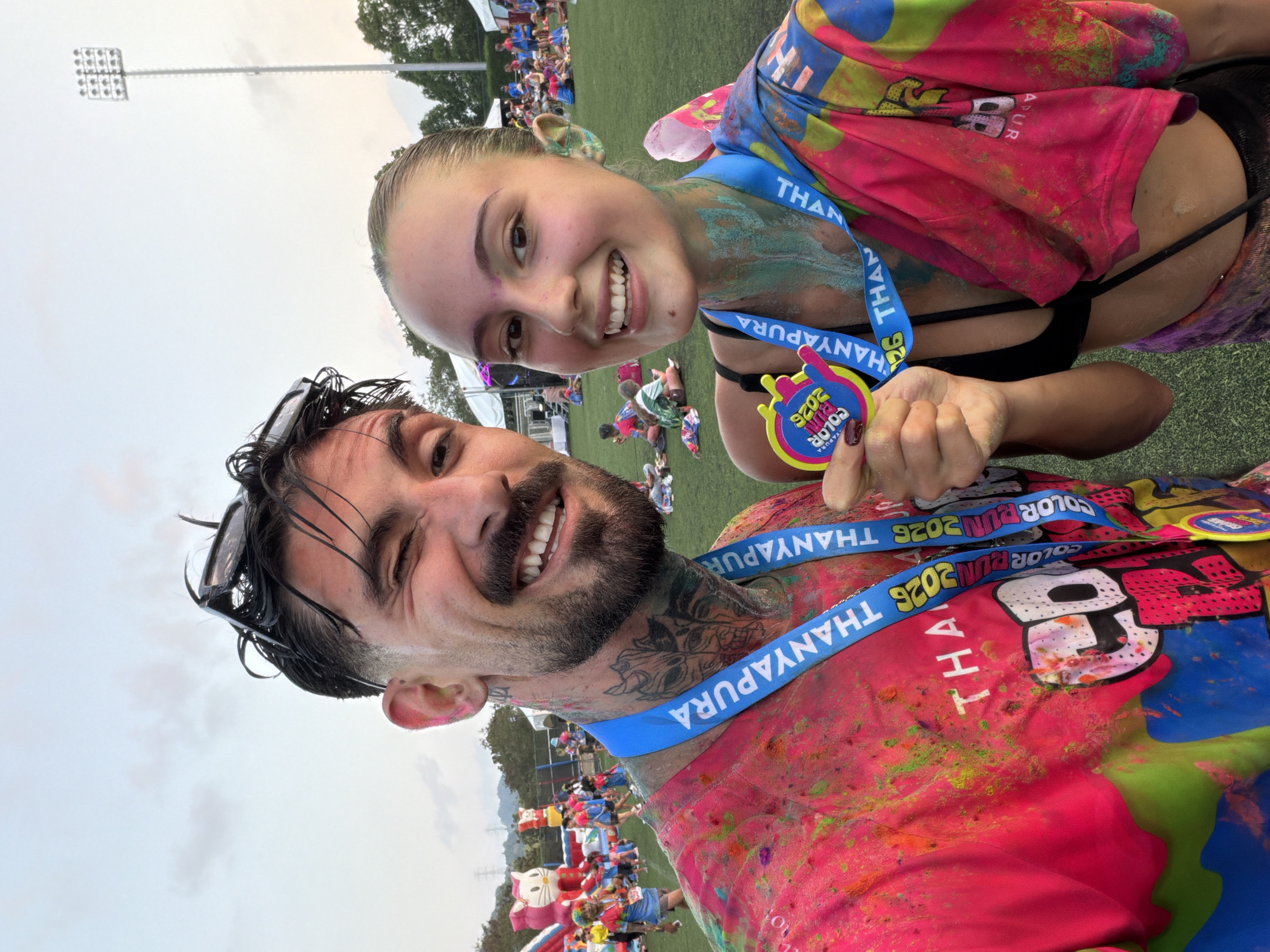 A selfie-style shot shows a smiling man and woman, presumably a couple, after participating in a "Color Fun" event. Both are covered in colored powder, suggesting a color run or similar activity. The man has a beard, tattoos on his neck, and wet hair with sunglasses perched on his head. The woman has her hair pulled back and is holding a medal with the words "Color Run 2026" on it. They are both wearing medals that say "Thanyapura Color Run 2026".
In the background, there's a crowd of people, an inflatable cat and other decorations, indicating an organized event. In the far background are what appears to be distant hills, and there is a light pole visible. The scene is set outdoors on a grassy field.
Based on the wording on the medals and the "Thanyapura" text, the event took place in Thanyapura, which is located in the city of Thep Krasatti, Thailand.